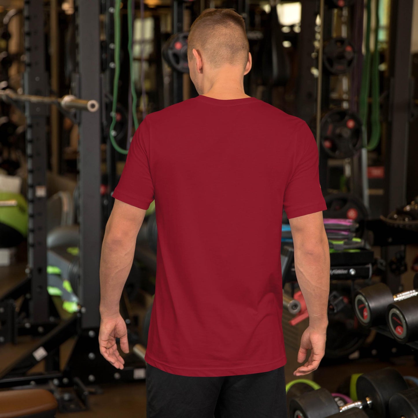 A man wearing a plain red Mambí t-shirt and black shorts stands with his back to the camera in a gym, surrounded by various exercise equipment and weights.