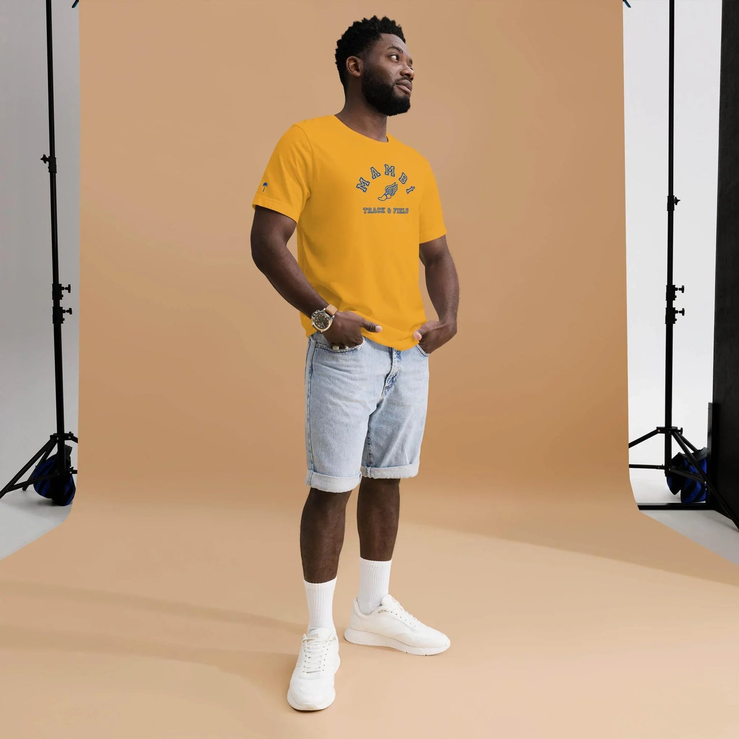A man stands in a photography studio with a beige backdrop, wearing Printfuls unisex Falcon Varsity Track & Field Tee with royal print, light denim shorts, white socks, and white sneakers—blending comfort and classic style.