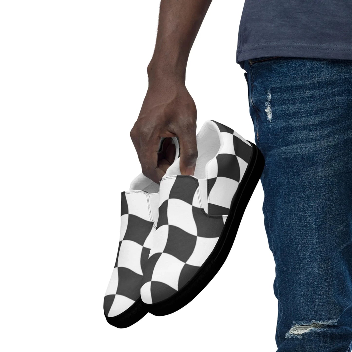A person wearing blue ripped jeans holds a pair of Mambí slip-on shoes with a black and white checkered pattern against a white background.