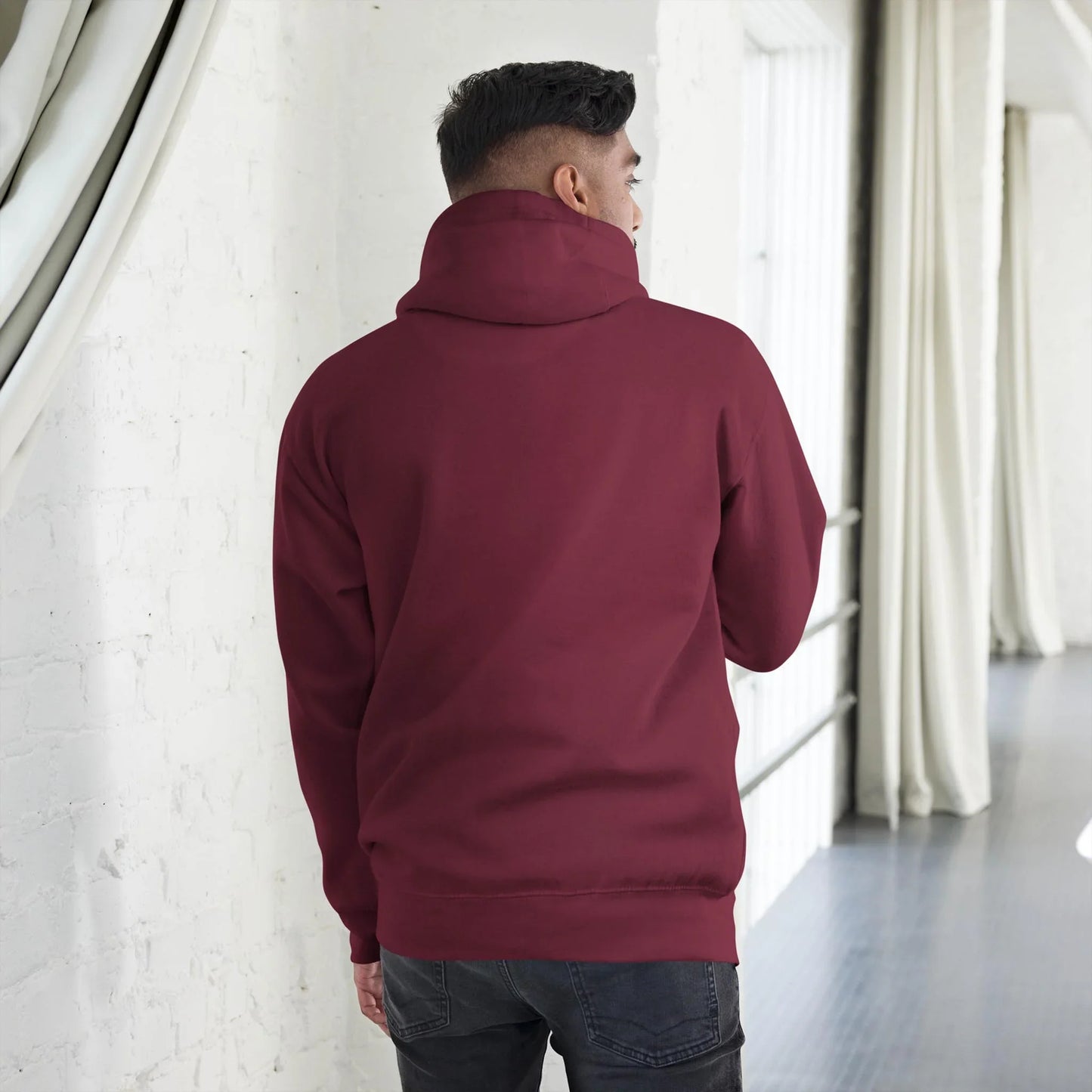 A person with short dark hair, seen from behind, is wearing a maroon Mambí hoodie and dark jeans, standing in a bright, modern room with white walls and long curtains.