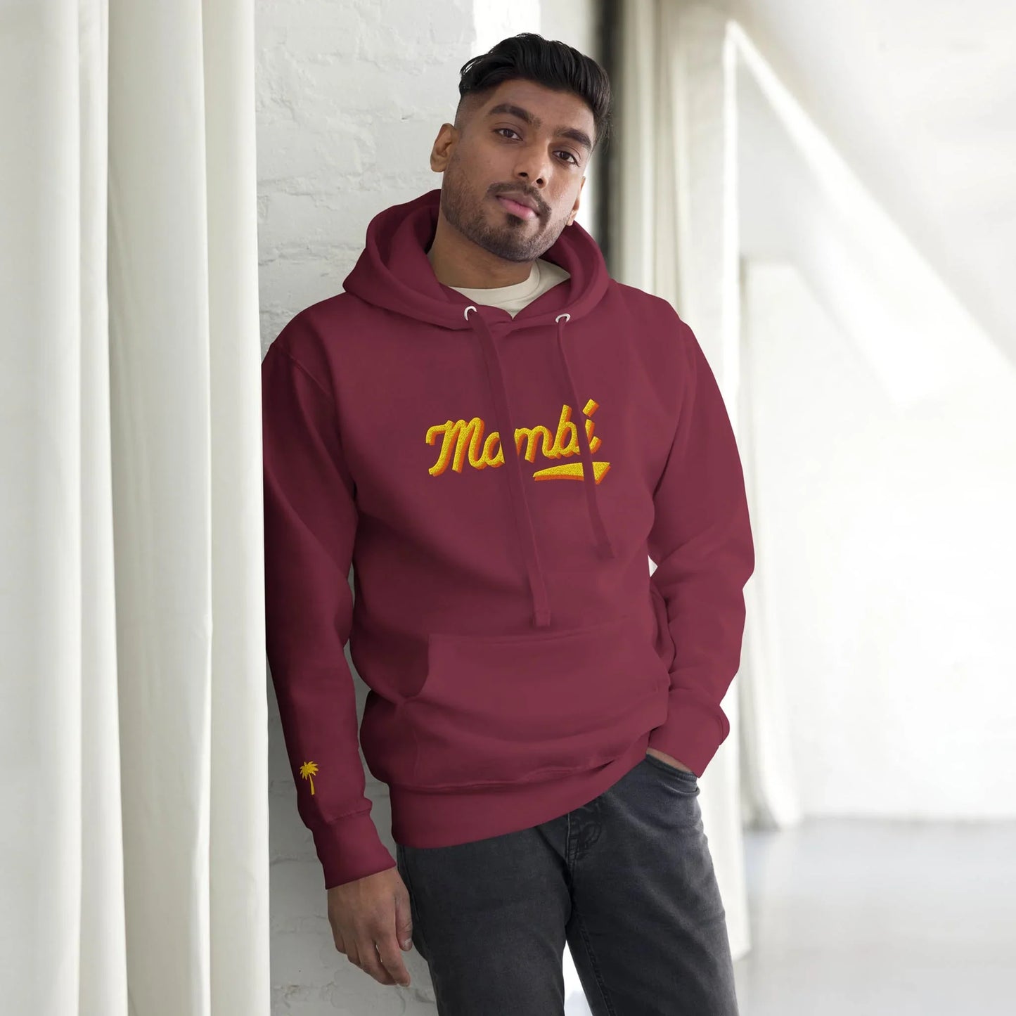 A man in a maroon Mambí hoodie with Mumbai written in yellow stands indoors, leaning against a white wall with curtains, looking at the camera with a relaxed expression.