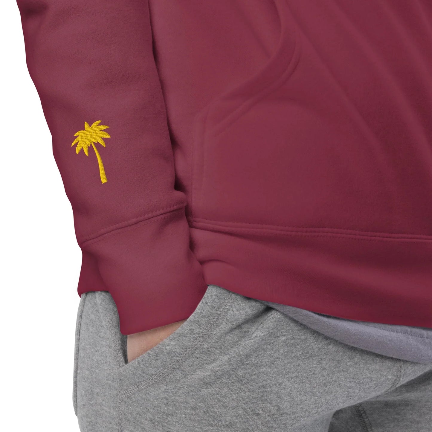 A person wearing a maroon Mambí sweatshirt with a small yellow palm tree embroidered on the sleeve and gray sweatpants, with one hand in their pocket.