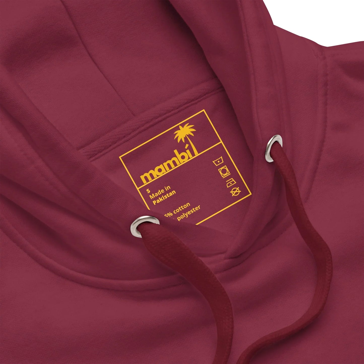 Close-up of a maroon hoodie with a yellow label inside the hood displaying the Mambí brand, palm tree logo, care symbols, and text stating Made in Pakistan, 80% cotton, 20% polyester.