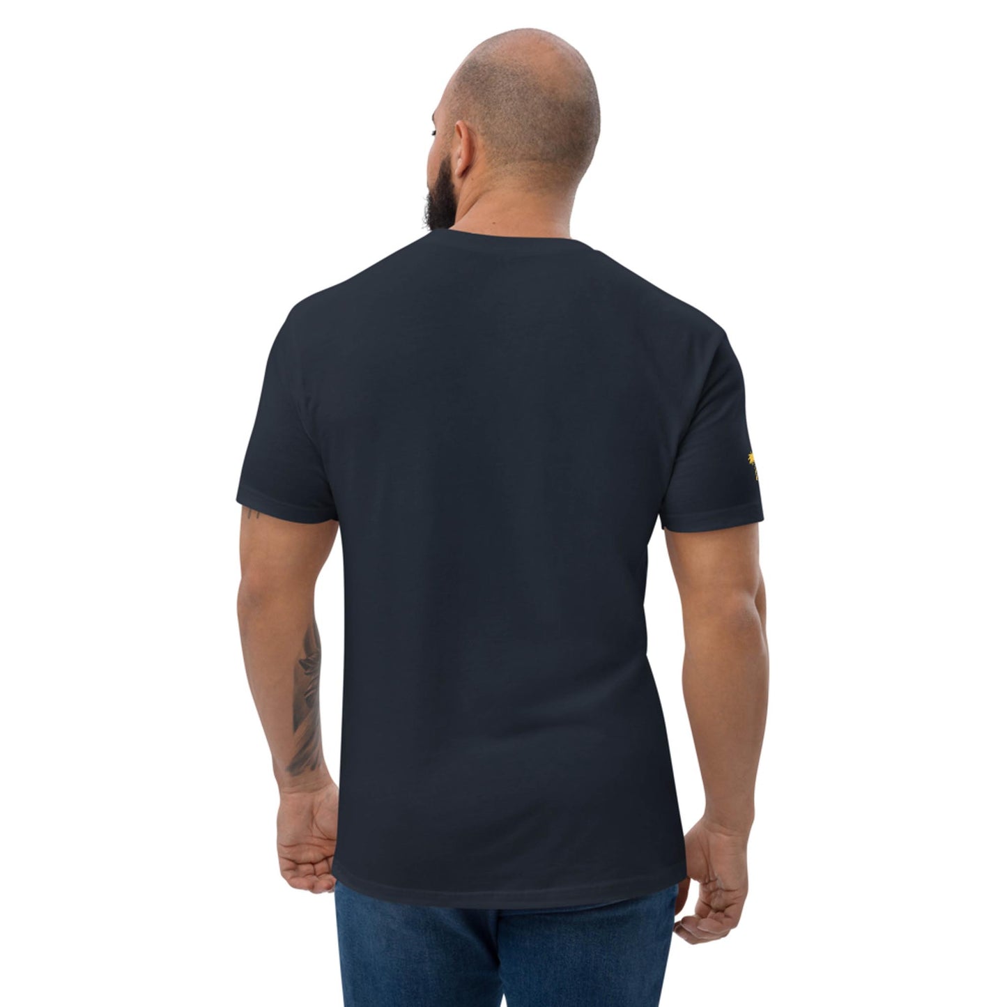A man with a shaved head and beard stands with his back to the camera, wearing a plain black Mambí t-shirt and blue jeans. His right arm features a visible tattoo.