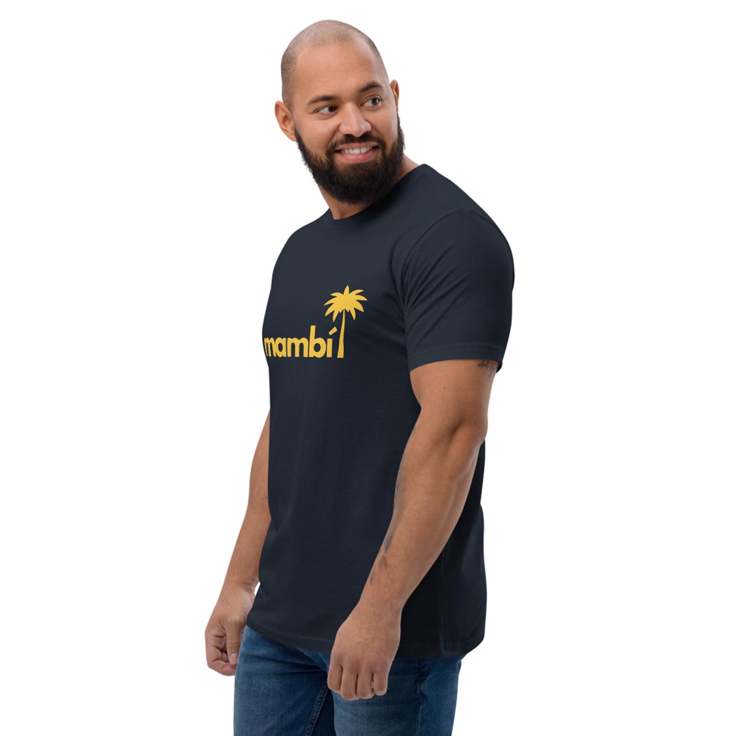A man with a beard and shaved head wears a dark Mambí t-shirt featuring a yellow palm tree graphic. He stands, smiling, and glances to his left against a plain white background.