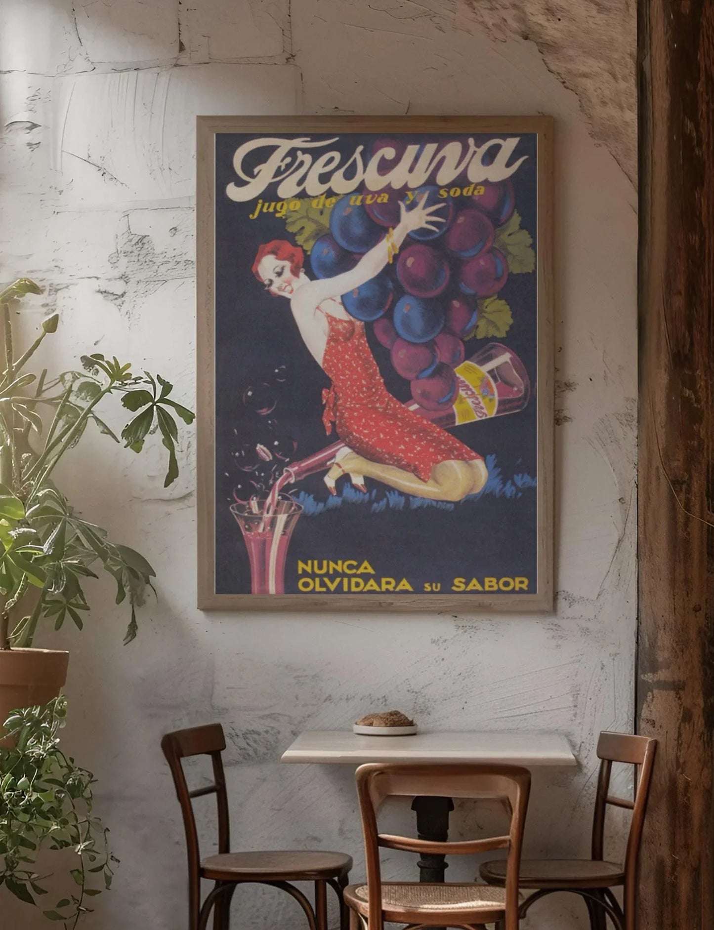 A vintage Mambí poster of a woman in a red dress holding large grapes hangs on a textured wall above a small café table with two chairs, a potted plant, and a pastry on a plate.