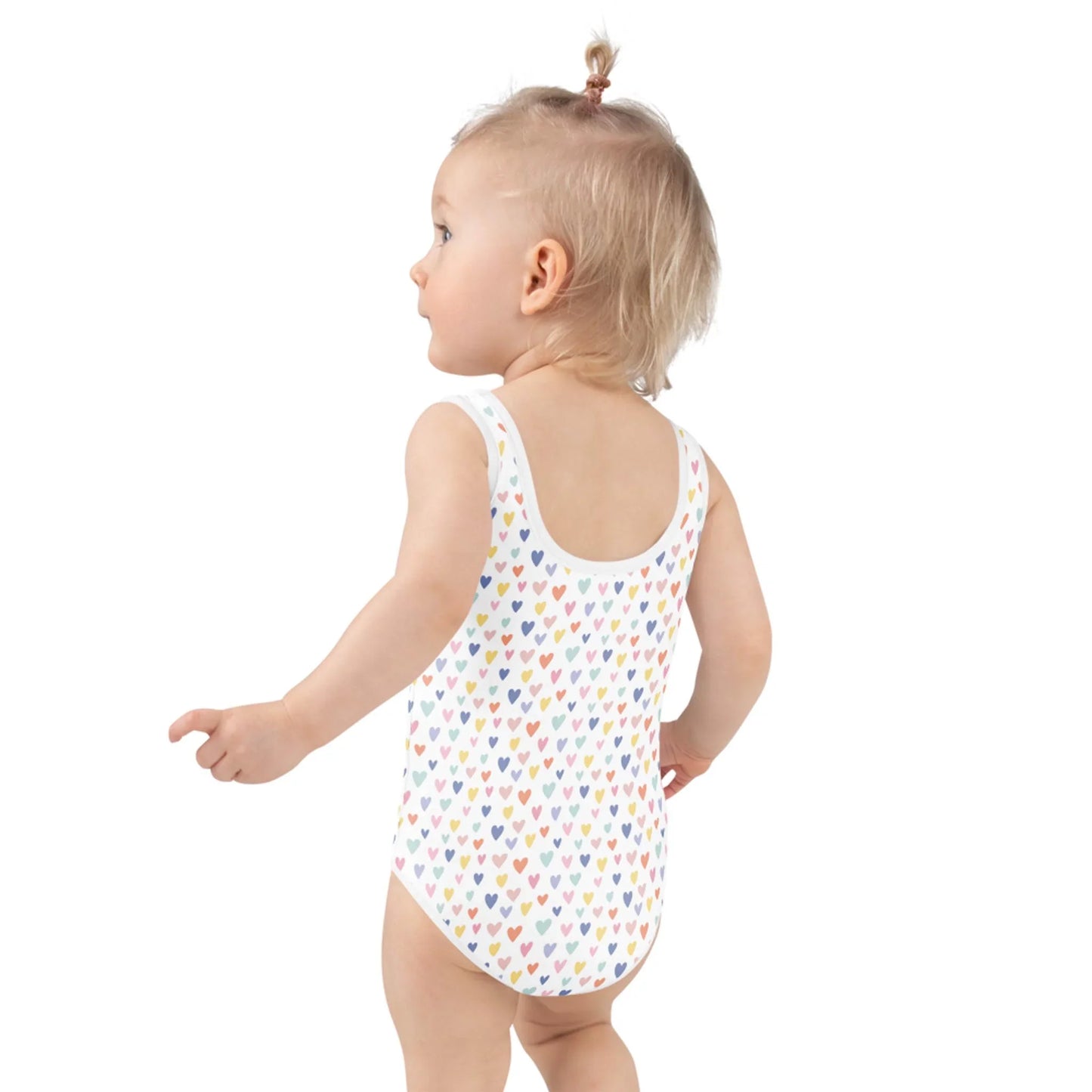 A toddler with a blonde ponytail wears the Printful Gemma One-Piece Swimsuit for toddlers, featuring colorful hearts and UPF sun protection, standing and facing slightly away from the camera.