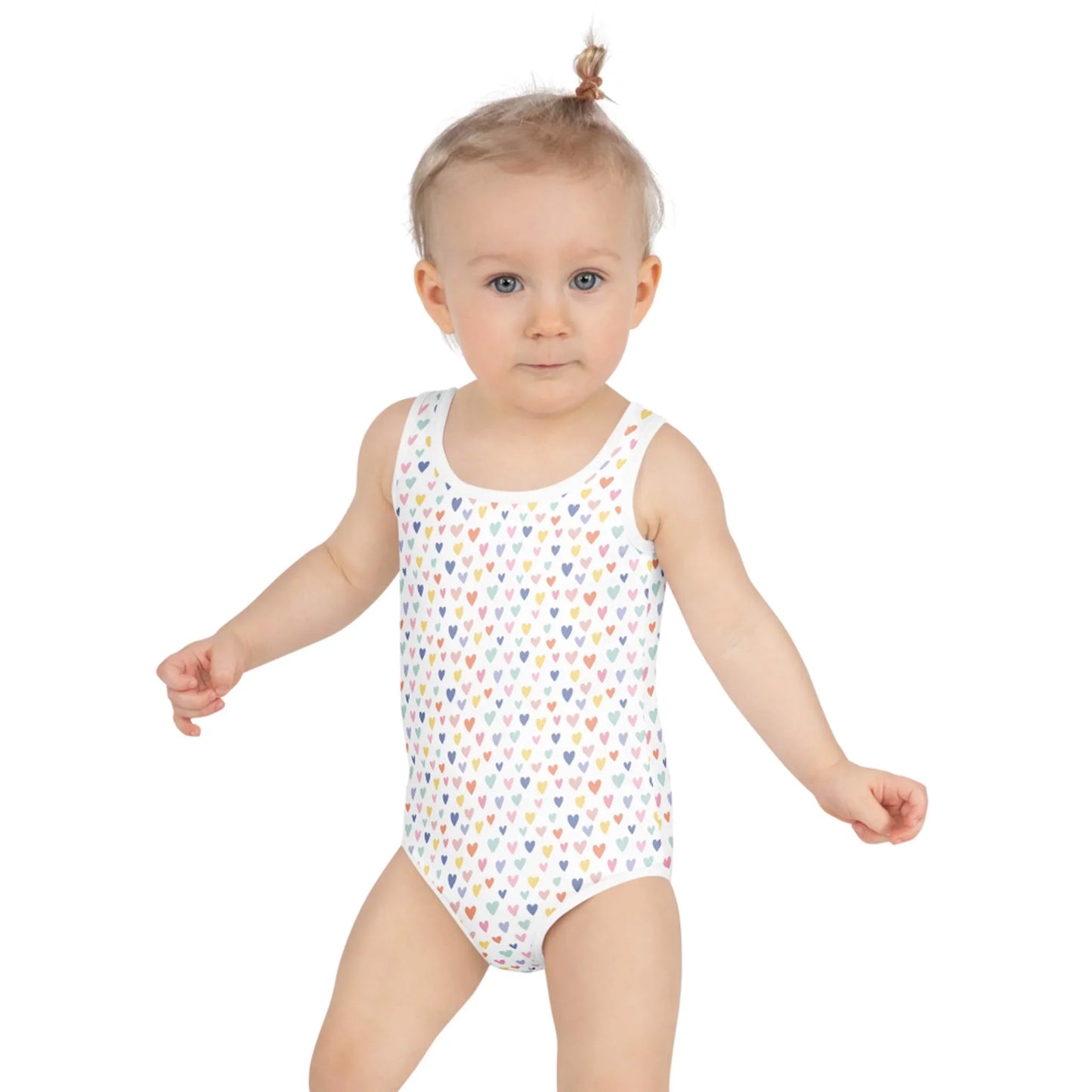 A light-haired toddler in a small topknot models Printful’s Gemma One-Piece Swimsuit for toddlers, featuring multicolored heart patterns and UPF sun protection, against a plain white background.