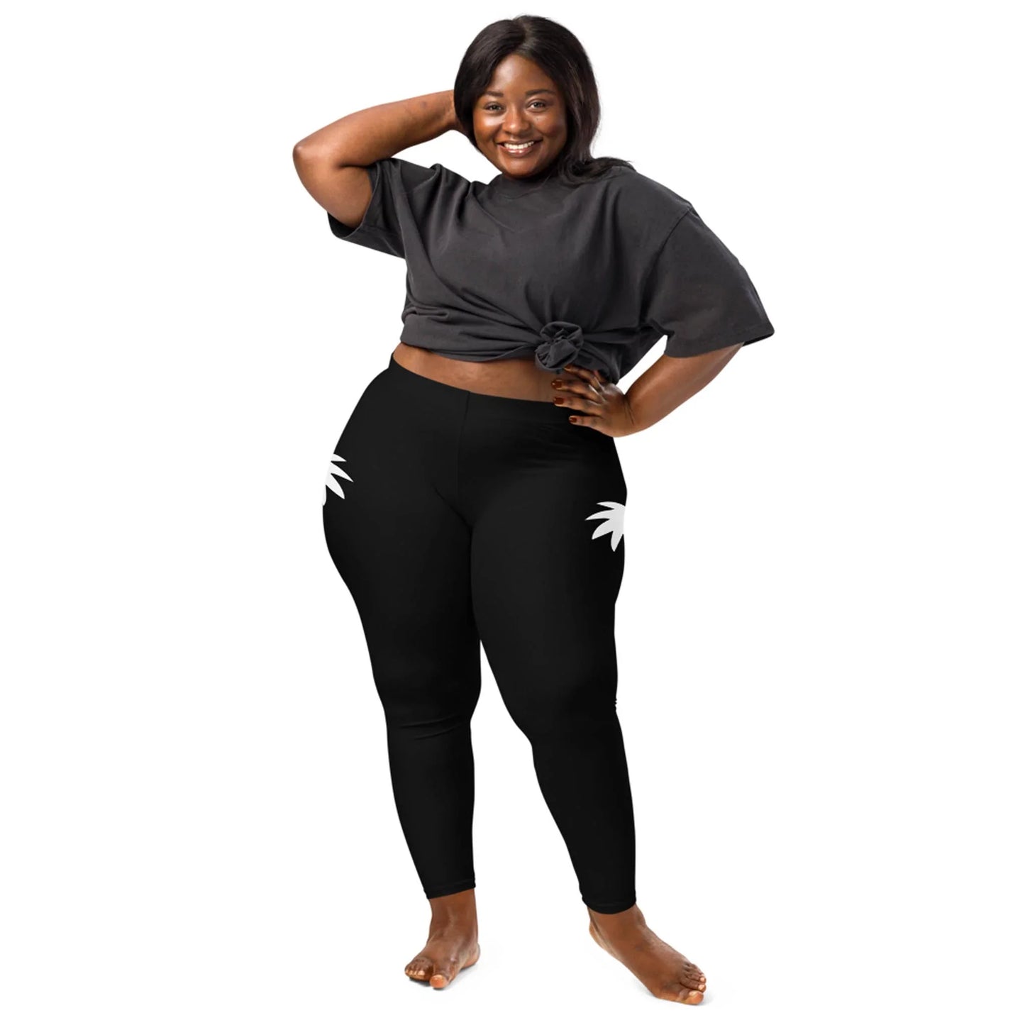 A smiling woman with dark skin and long hair poses barefoot, wearing a tied black Mambí t-shirt and black leggings with white palm leaf designs on the hips, standing against a white background.