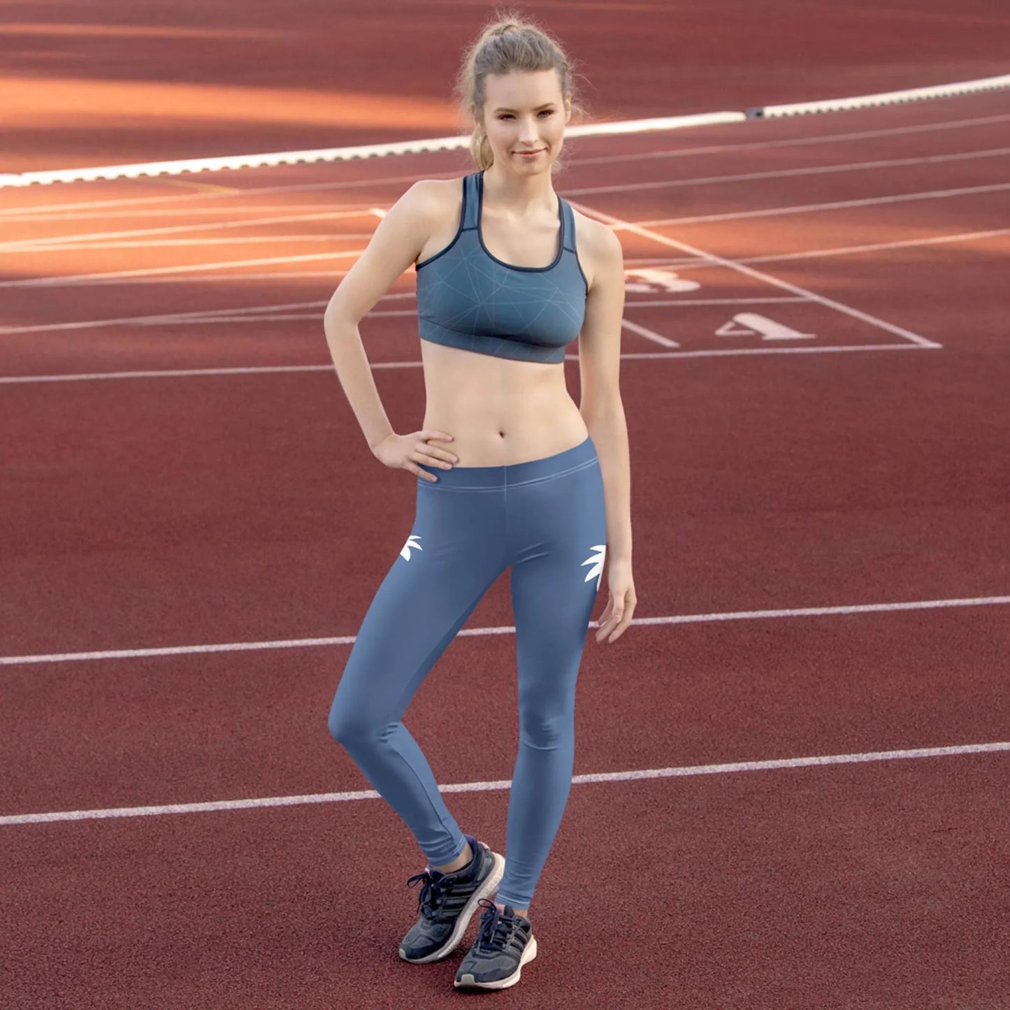 A woman in blue Mambí athletic wear stands on a red running track, posing confidently with one hand on her hip and the other relaxed by her side. She wears a sports bra, leggings, and running shoes, with track lines visible in the background.
