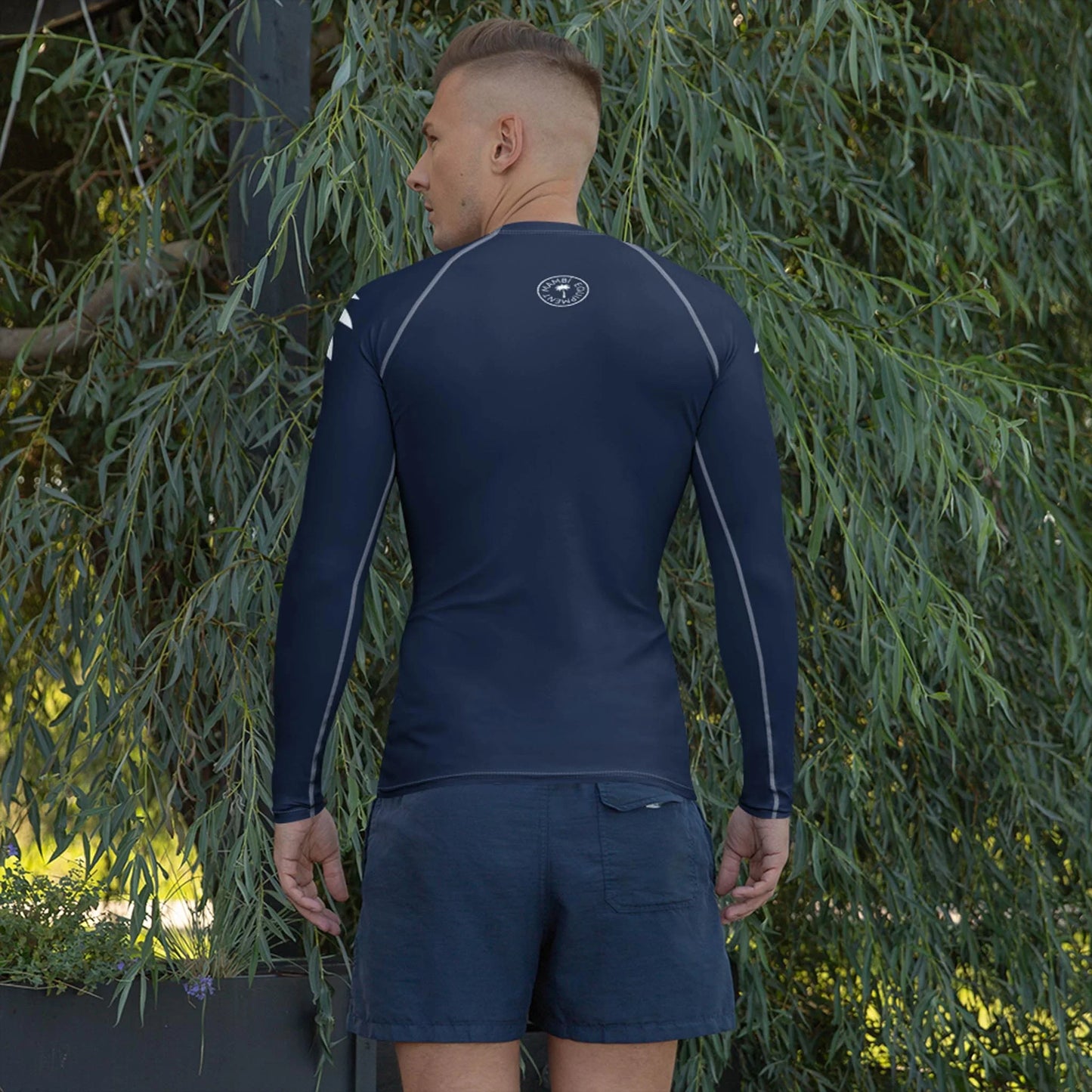 A man with short hair stands outdoors, facing away, wearing a navy blue Mambí long-sleeve athletic shirt and matching shorts. Lush green foliage fills the background.