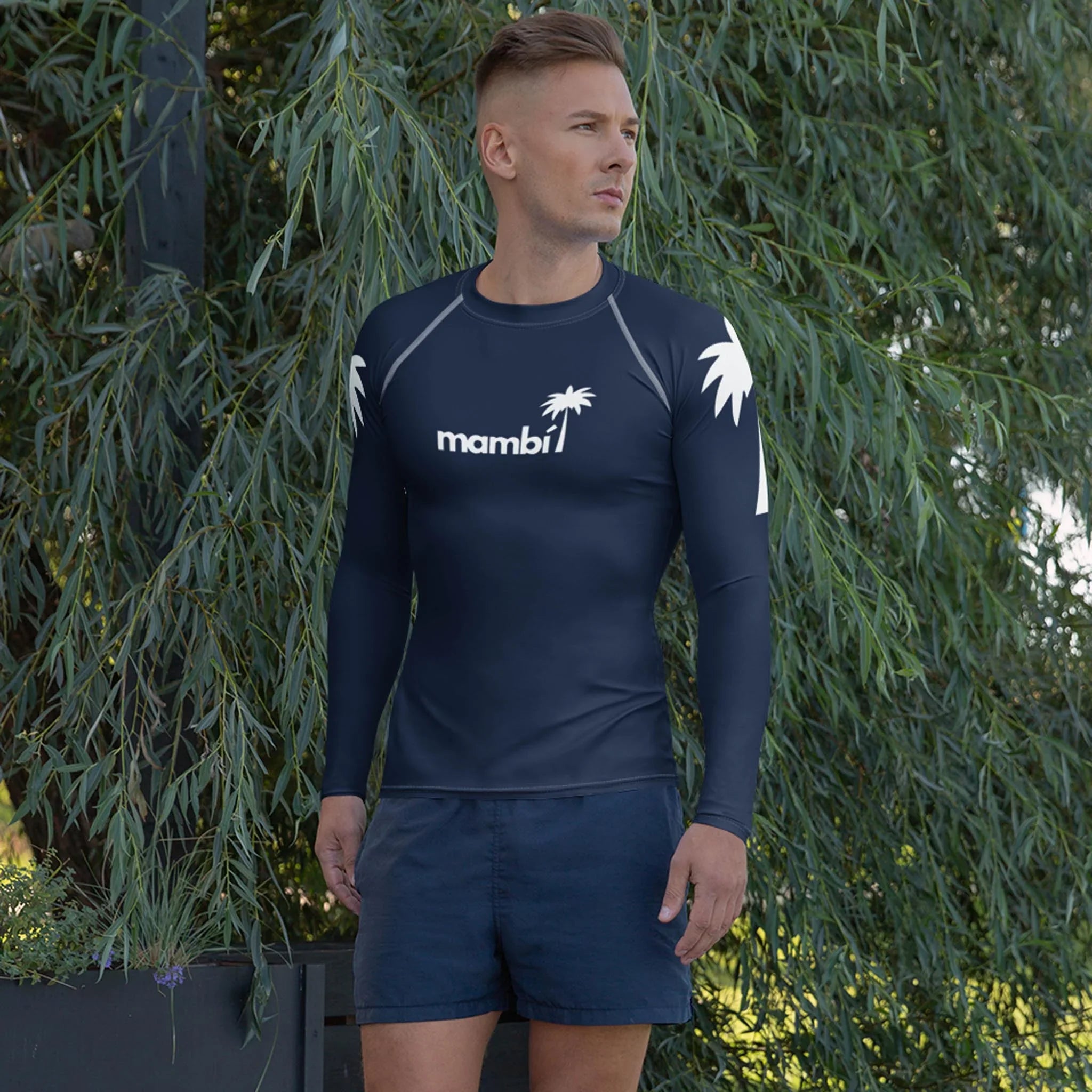A man stands outdoors by leafy greenery, wearing the Printful Hasselhoff Classic Long-Sleeve Rash Guard for men with UPF 50+ and a white palm tree mambi1 graphic on the chest, paired with navy shorts—ideal for watersports.