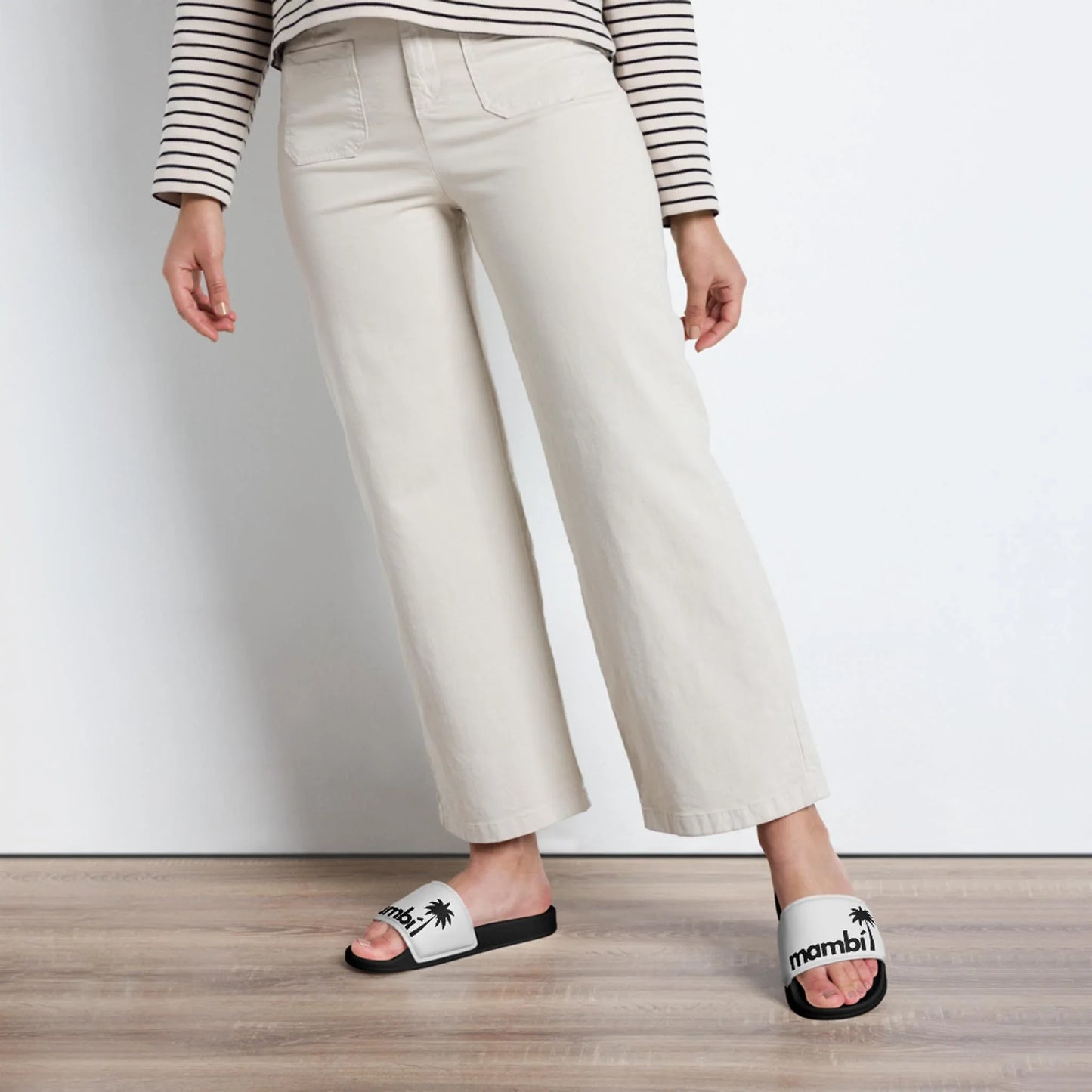 A person wearing a striped long-sleeve shirt, light beige pants, and white Mambí slide sandals with black soles stands on a wooden floor against a white background.