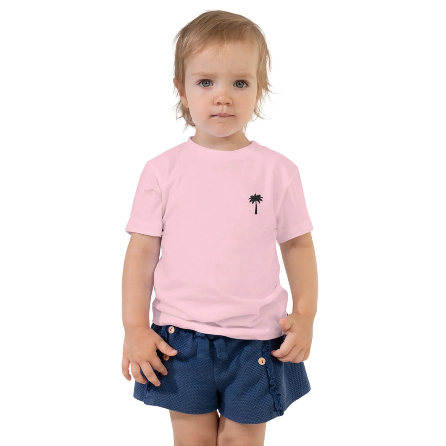A young child with light hair wears a light pink Mambí t-shirt featuring a small black palm tree graphic on the chest and blue shorts with buttons, standing against a plain white background.