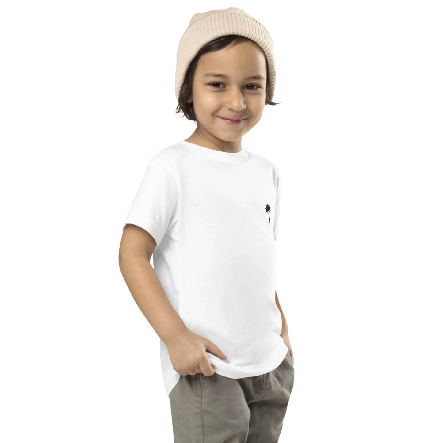 A young child wearing a Mambí white t-shirt, beige knit beanie, and khaki pants stands with hands in pockets, smiling at the camera against a plain white background.