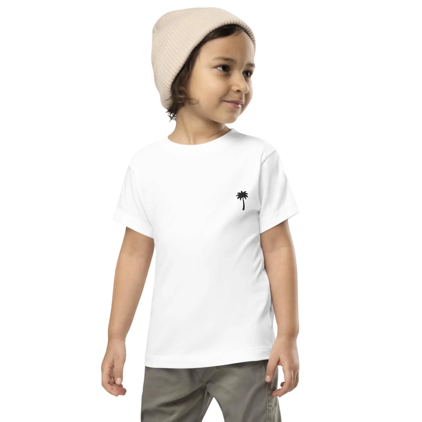 A young child wearing a beige knit beanie, white t-shirt with a small black palm tree graphic on the chest, and khaki pants, smiling and looking to the side against a plain background—styled by Mambí.