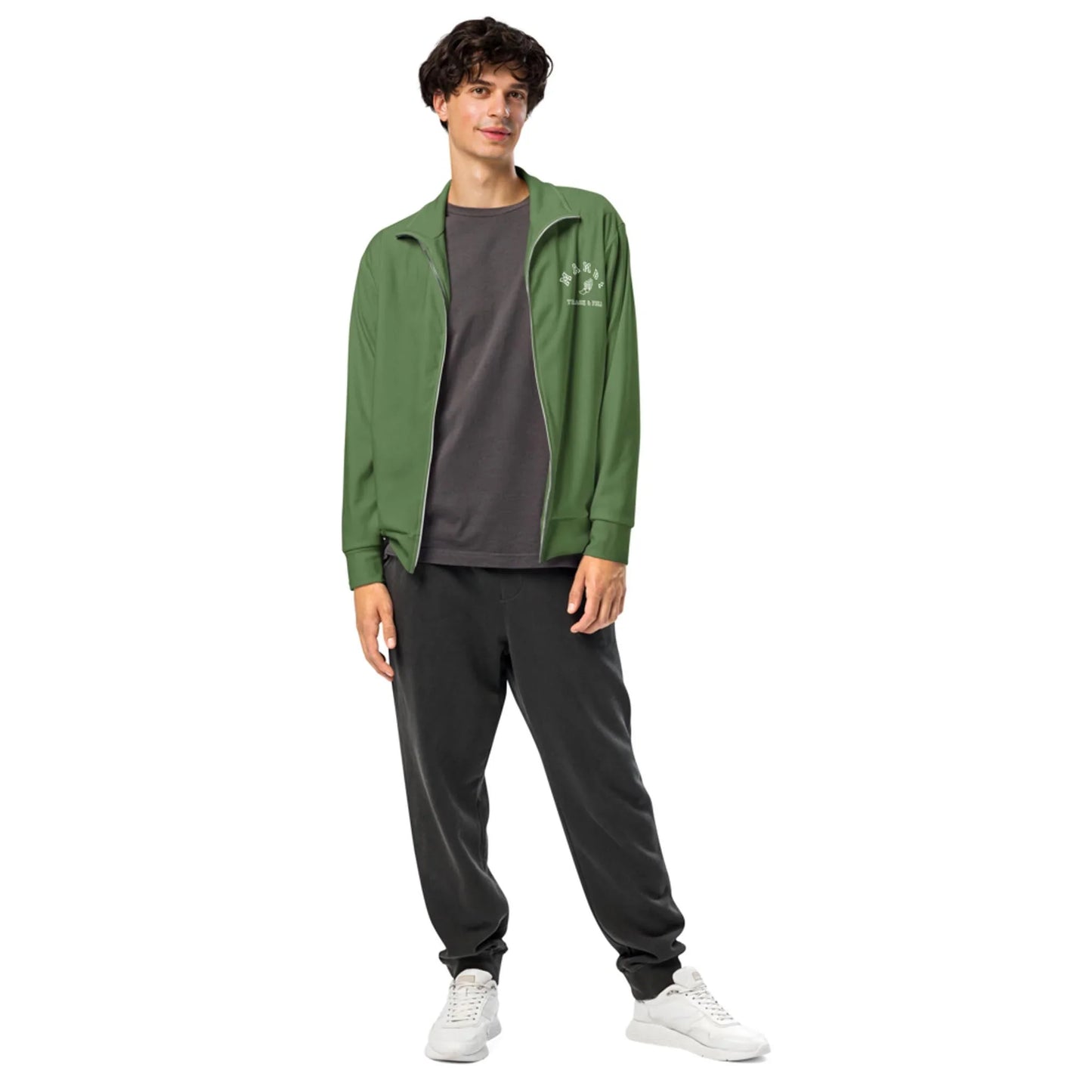A young man stands casually, wearing a green Mambí zip-up jacket over a dark gray t-shirt, black jogger pants, and white sneakers. He is smiling and looking at the camera against a plain white background.