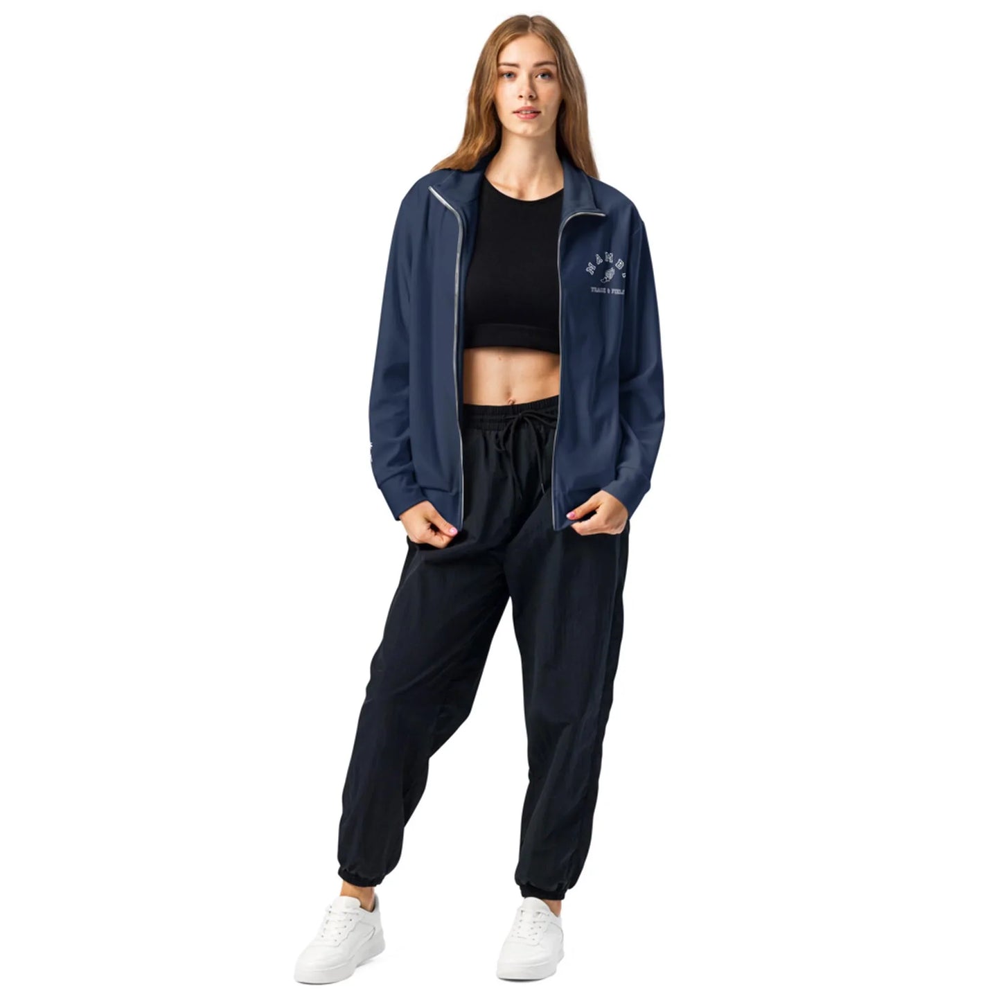 A woman stands wearing a Mambí navy zip-up jacket over a black crop top, black jogger pants, and white sneakers, posing with one hand in her pocket against a white background.