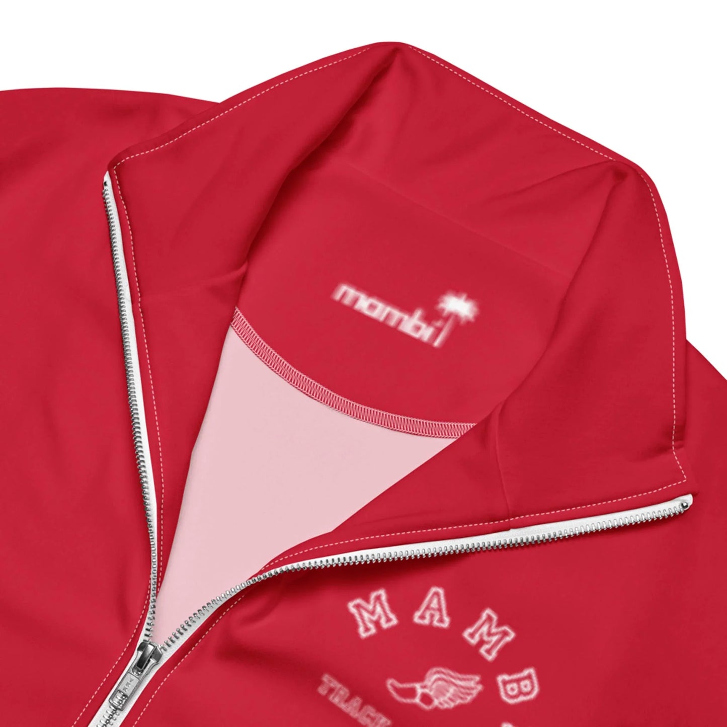 Close-up of a red zip-up jacket with white embroidery reading Mambí and a soccer ball with wings. Inside the collar, Mambí and a palm tree are printed in white. The zipper is partially open, revealing a light pink lining.