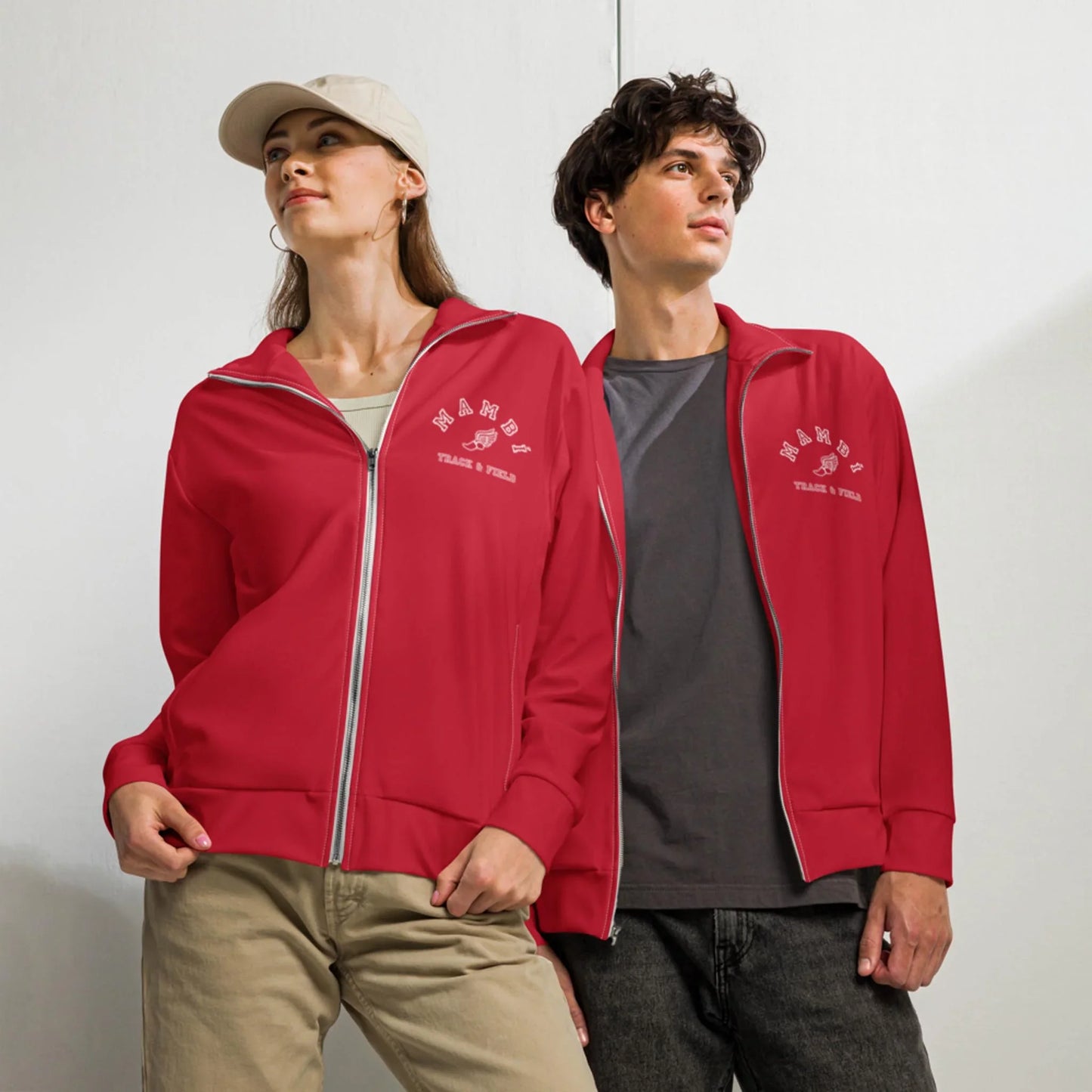 Two people pose together in matching red Mambí zip-up jackets with white text on the left chest. The person on the left wears a beige cap and hoop earrings, while the person on the right looks sideways against a plain white wall.