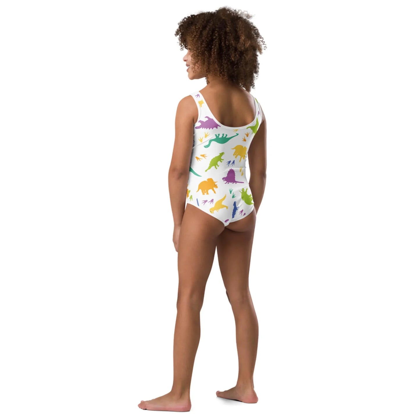A young girl stands barefoot facing away, wearing a white Mambí one-piece swimsuit with colorful dinosaur patterns. She has curly hair and stands against a plain white background.