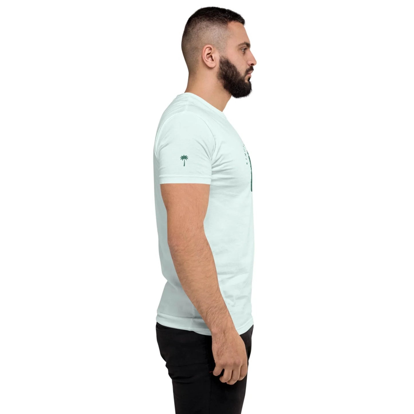 A man with a beard stands in profile, facing left, wearing a light blue Mambí T-shirt and black pants against a plain white background.
