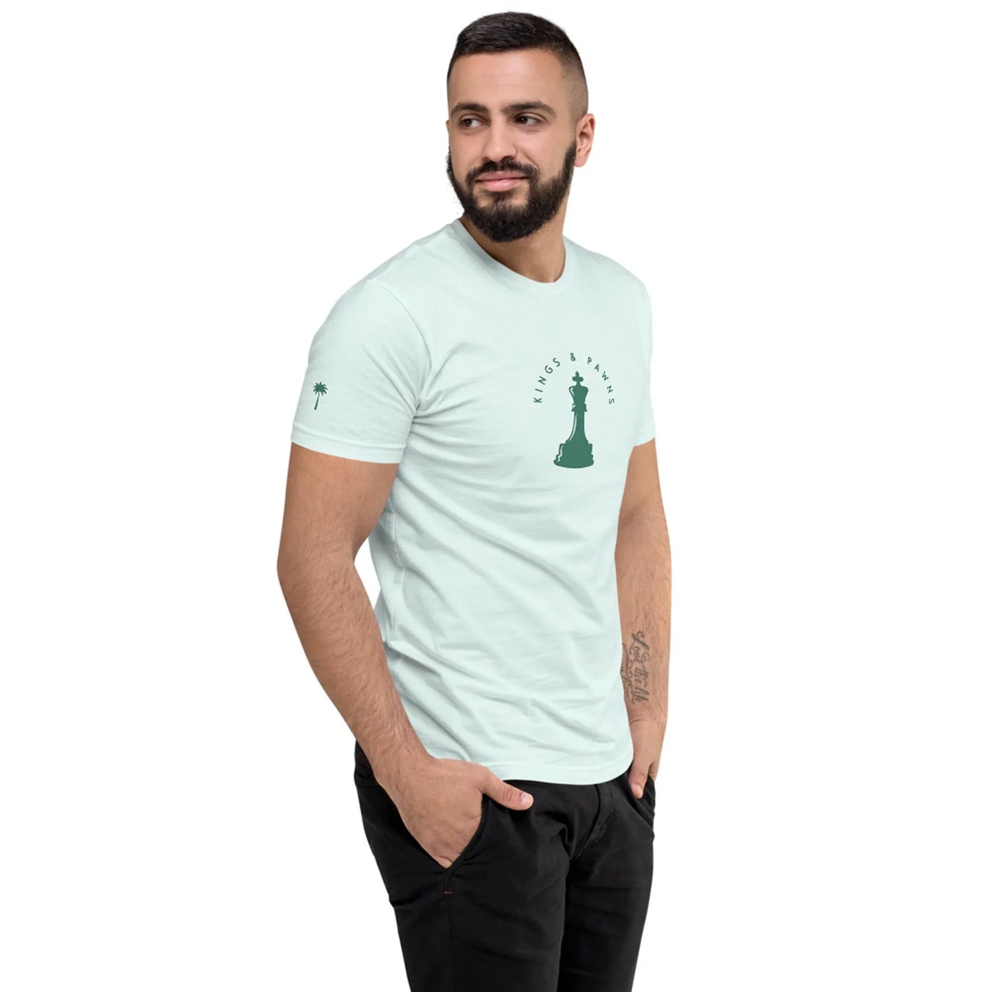 A man with short dark hair and a beard wears a Mambí t-shirt featuring a chess king graphic and the words “King’s Pawn” on the chest. He stands with hands in his pockets, looking to the side against a white background.