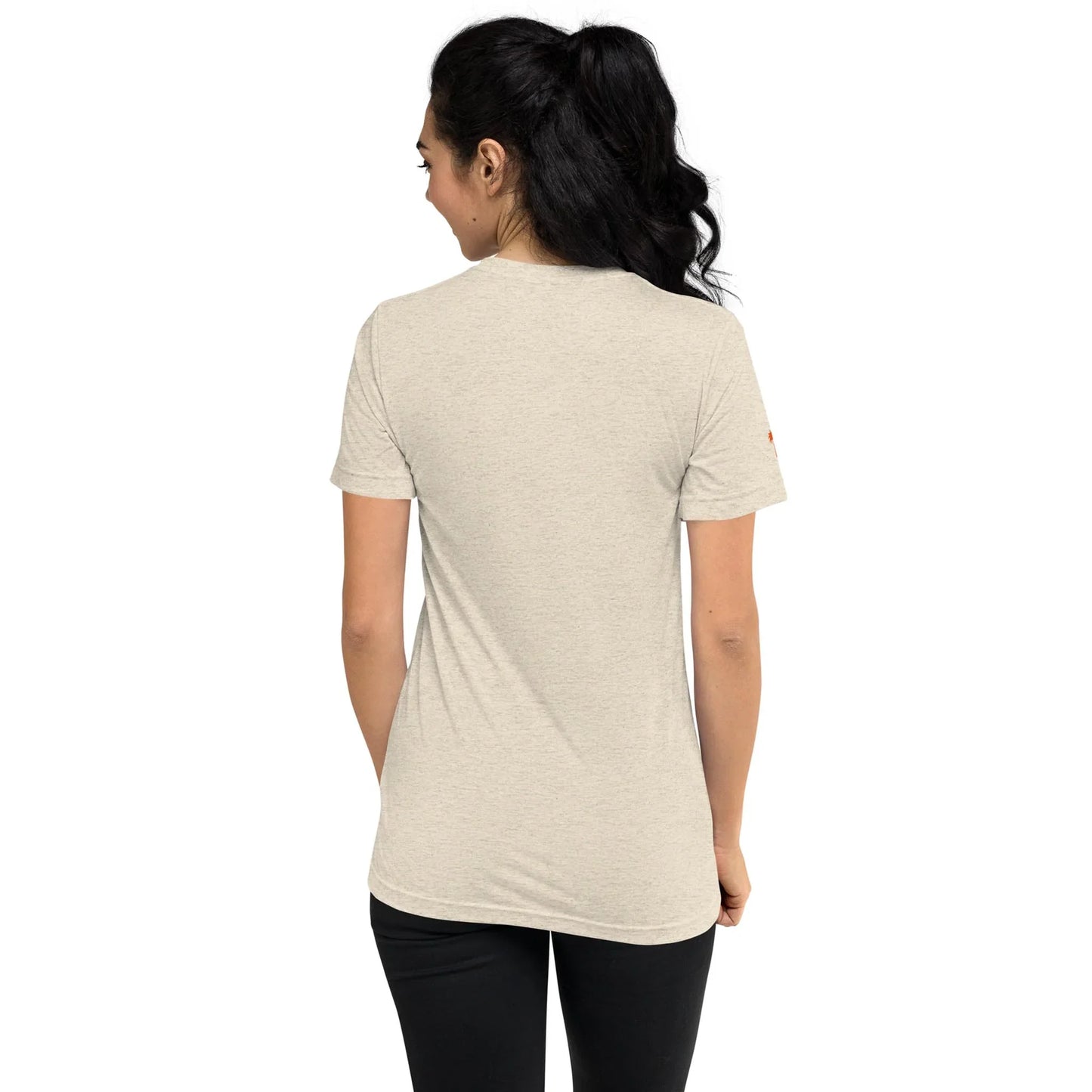 A woman with long dark hair in a ponytail stands with her back to the camera, wearing a light beige Mambí short-sleeve t-shirt and black pants against a plain white background.