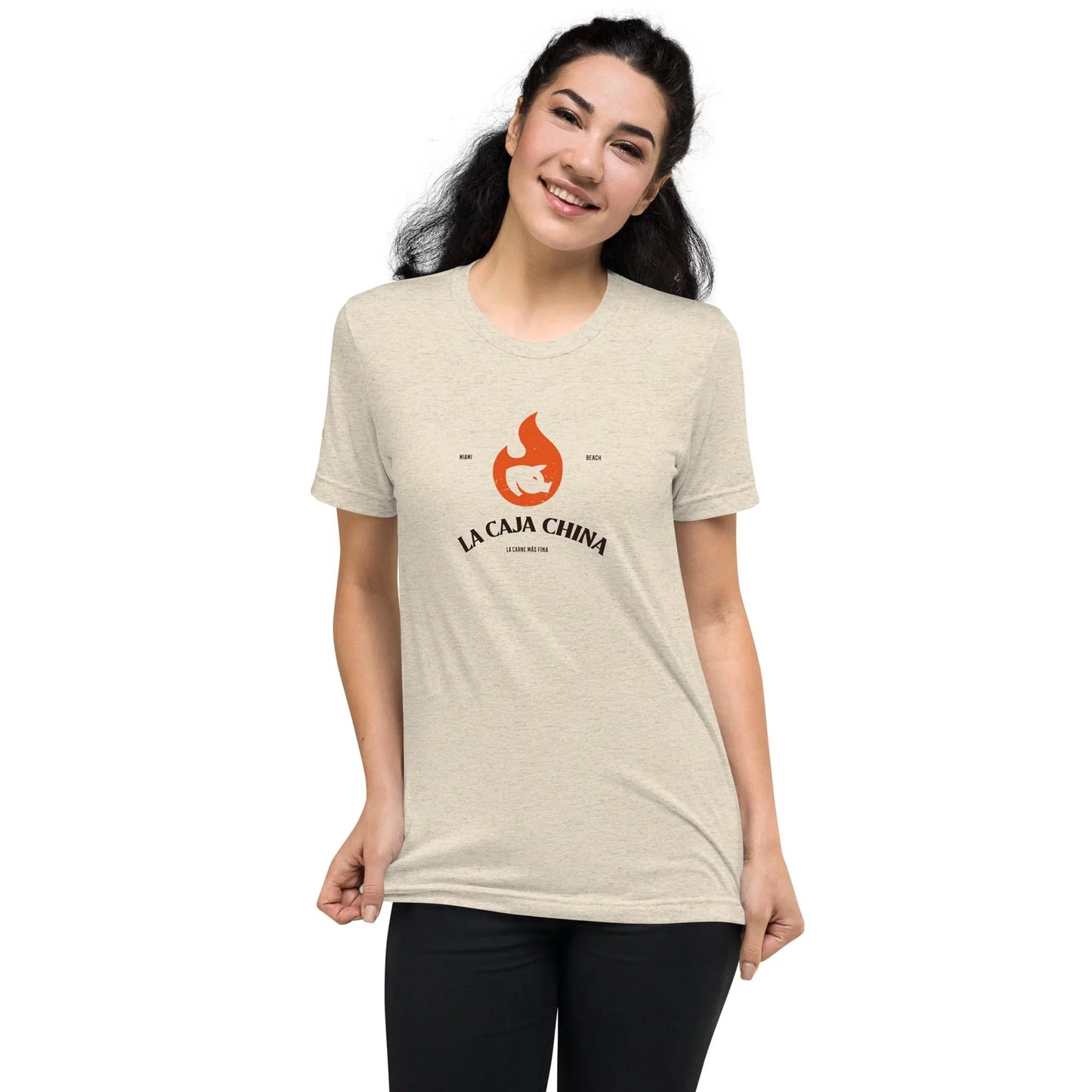 Smiling person with long dark hair wearing a light-colored t-shirt featuring an orange flame graphic, the text LA CAJA CHINA, and a subtle Mambí touch on the front. They are standing against a plain white background.