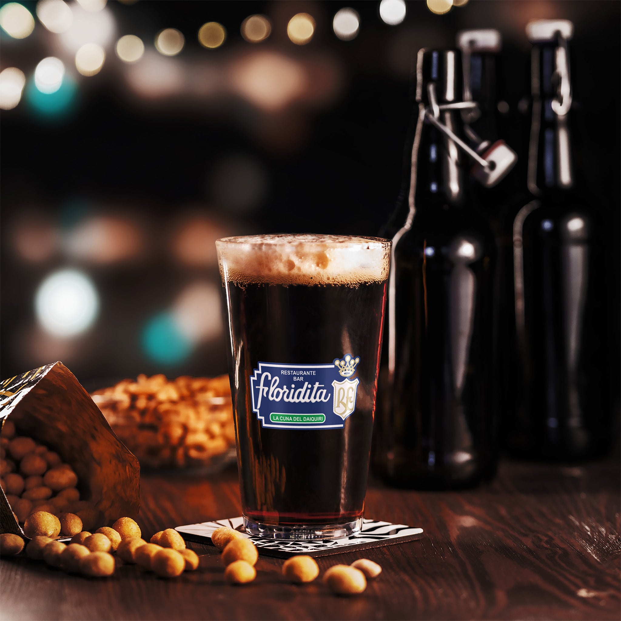 A glass of dark beer with a foamy head sits on a coaster on a wooden table beside two sealed bottles and a tipped-over container spilling peanuts. Blurred lights shine in the background.