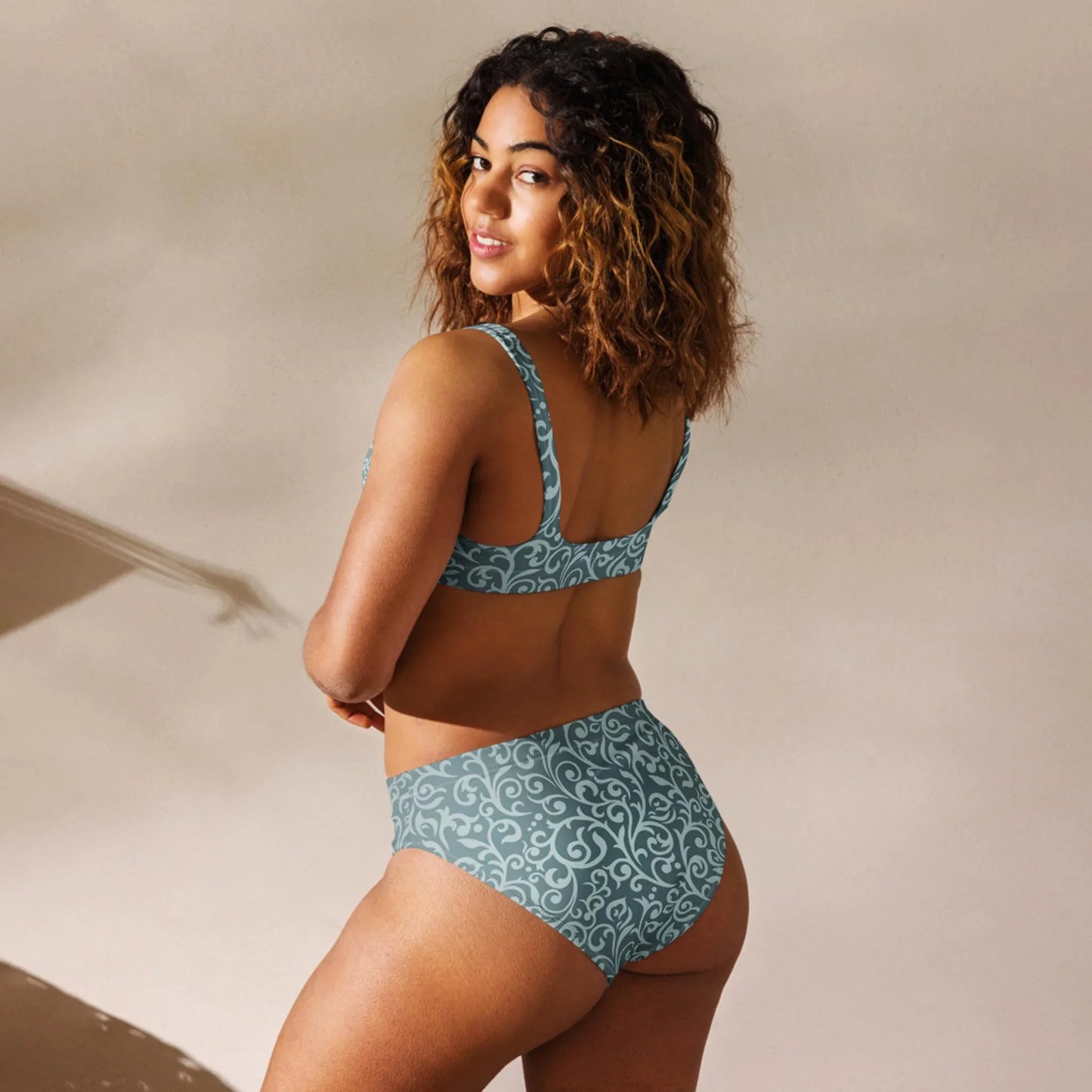 A woman with curly hair models the Printful Laurelia High-Waisted Bikini Swimsuit, featuring a matching light blue design with white swirls, standing sideways and glancing over her shoulder against a neutral background.