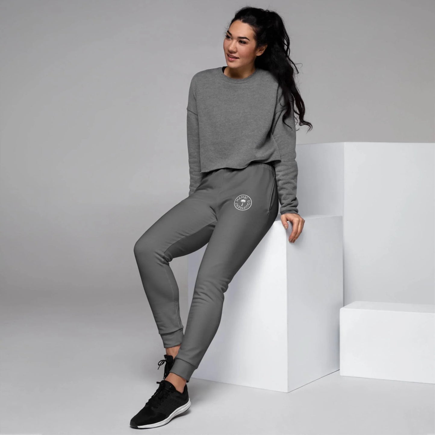 A woman with long dark hair, wearing a gray Mambí sweatshirt and gray joggers, sits on a white block in a minimal studio. She is smiling and dressed in black sneakers.