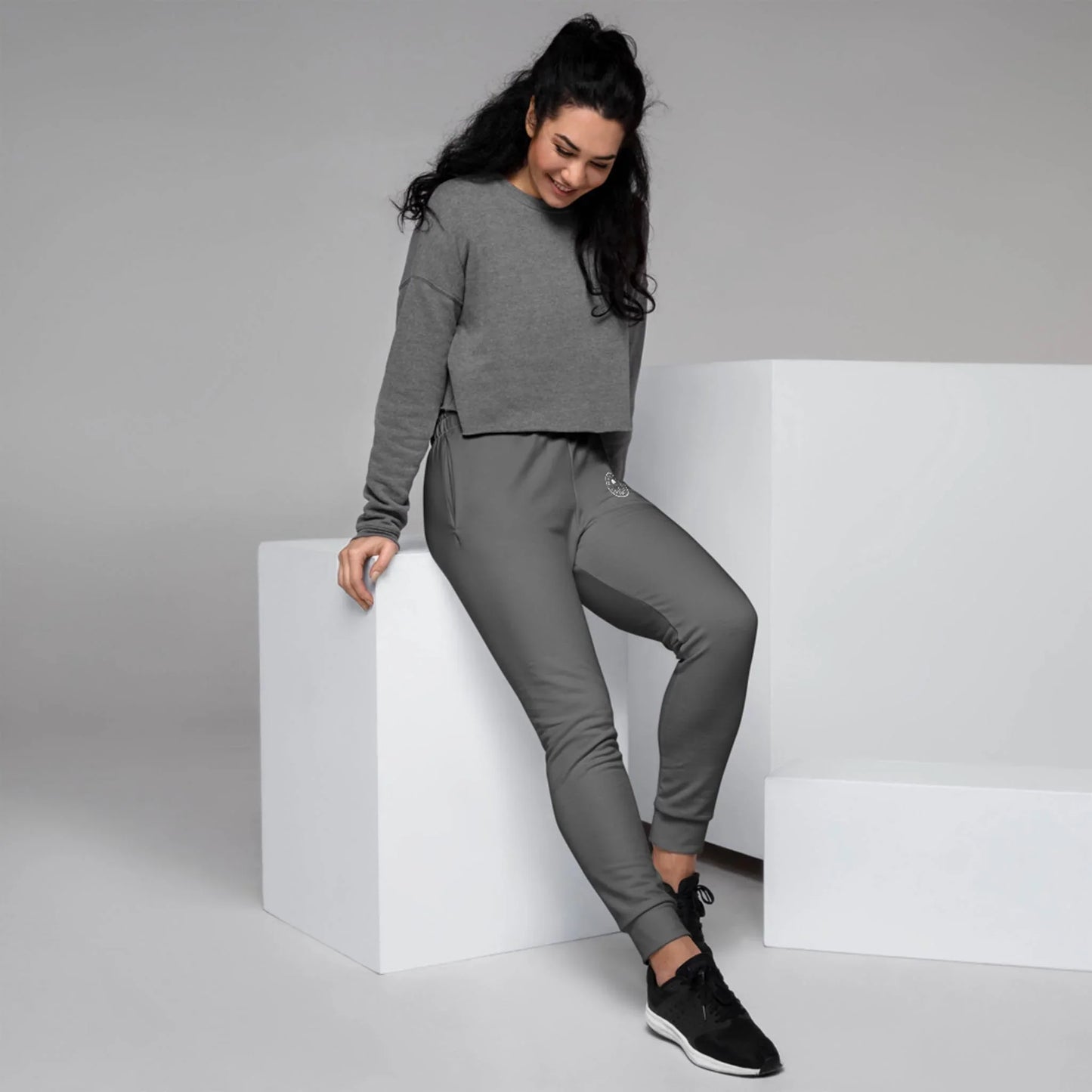 A woman with long dark hair wears a Mambí grey cropped sweatshirt and grey joggers, leaning casually against a white cube in a minimalist studio setting, smiling and looking down.