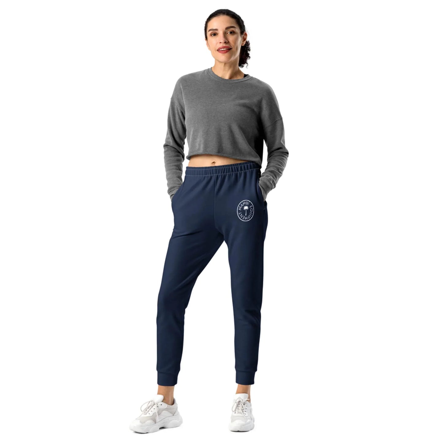 A woman stands against a white background wearing a gray Mambí long-sleeve crop top, navy jogger pants with a circular white logo on one leg, and white sneakers, her hands in her pockets as she smiles.