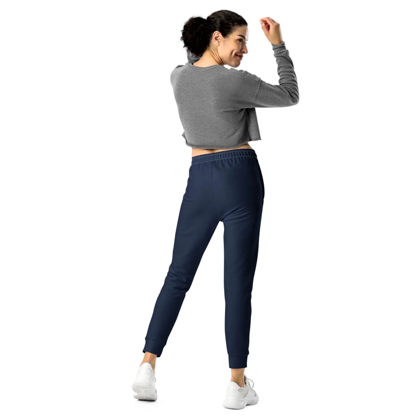 A woman with curly hair, wearing a gray long-sleeve crop top, navy jogger pants, and white sneakers from Mambí, stands facing away and smiles while looking back over her shoulder.