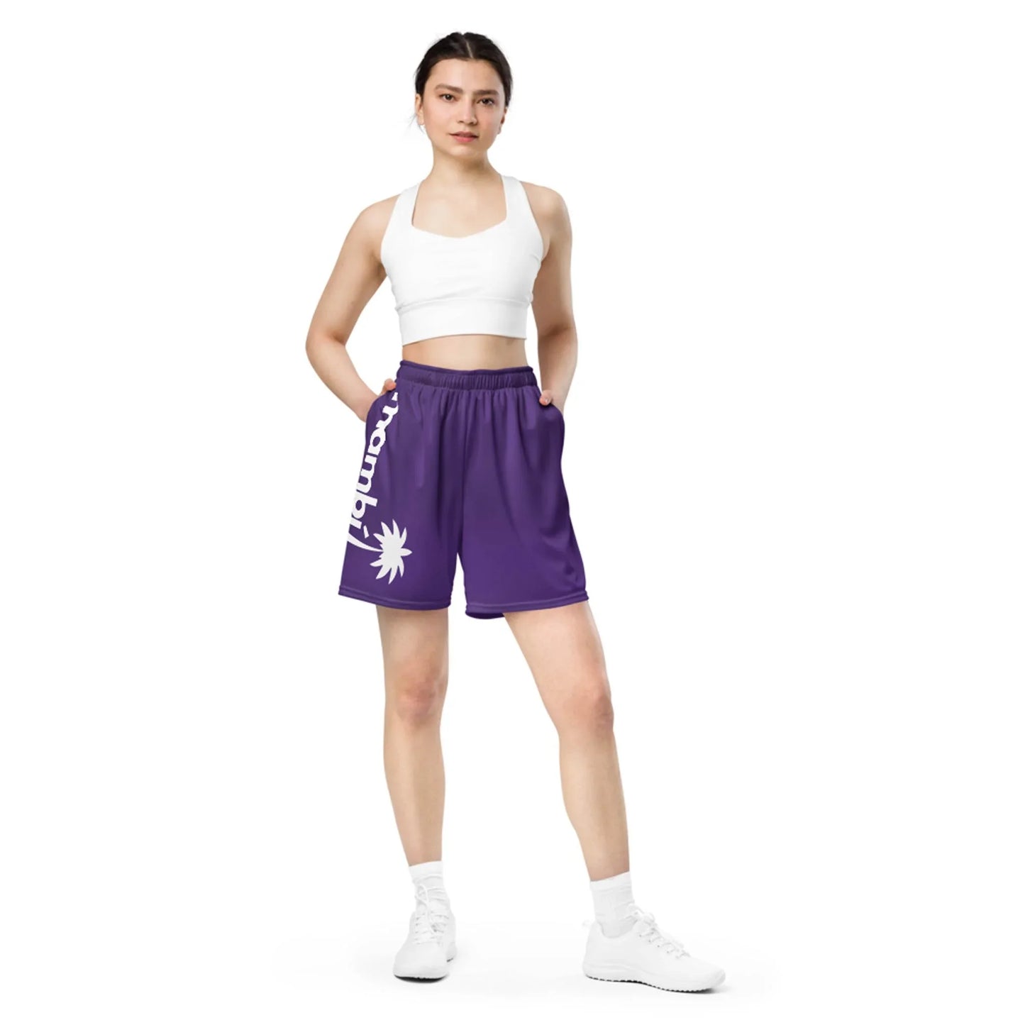 A person stands with hands in pockets, wearing a white crop top, Mambí purple athletic shorts featuring a white sun graphic and vertical text, and white sneakers against a plain white background.