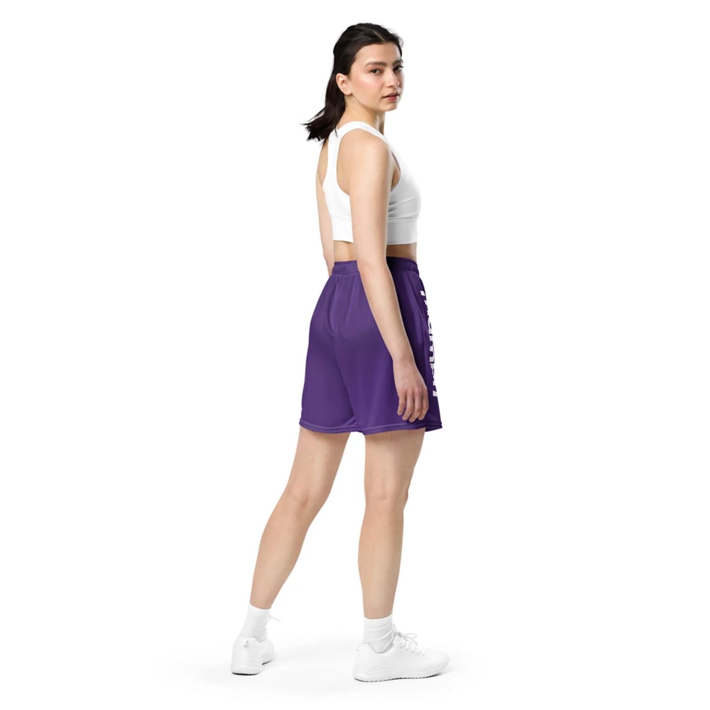 A woman stands turned slightly to the side, wearing a white Mambí crop top, purple athletic shorts, white socks, and white sneakers against a plain white background. She looks over her shoulder toward the camera.