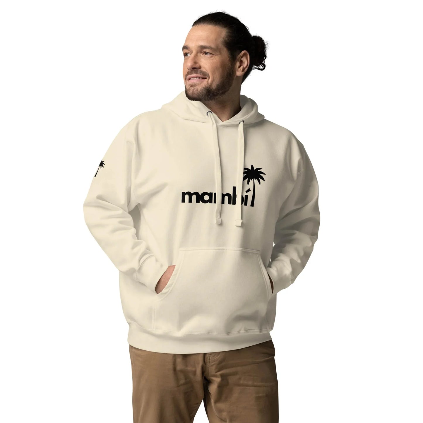 A man with a beard and tied-back hair wears a light hoodie featuring a Mambí miami and palm tree graphic, standing with hands in pockets, looking to the side and smiling against a plain white background.