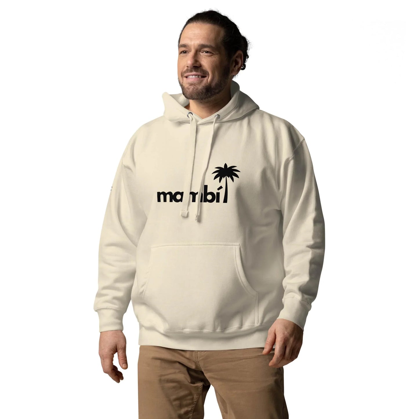 A man wearing a light-colored Mambí hoodie with a miami logo and palm tree design stands smiling against a plain white background. He has dark hair tied back and is also wearing tan pants.