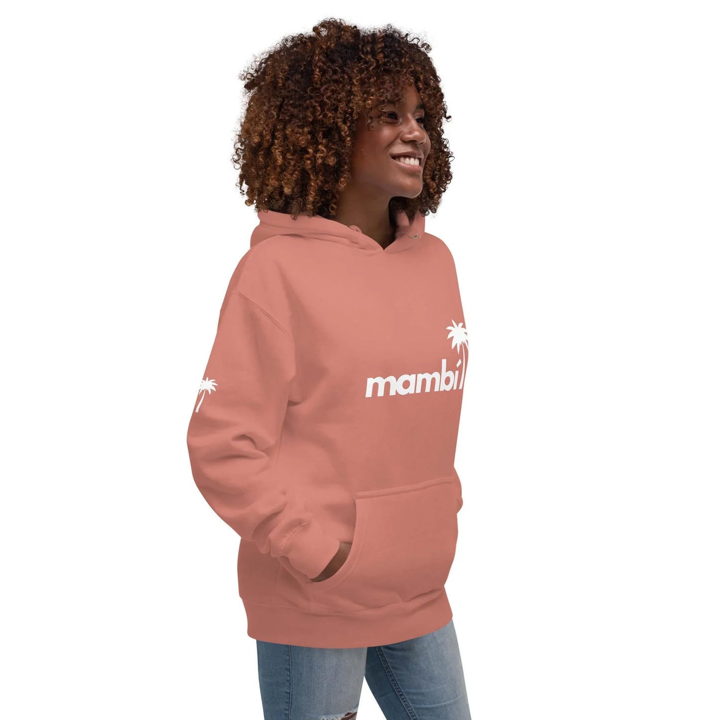 A smiling woman with curly hair wears a dusty pink Mambí hoodie featuring a white palm tree graphic on the front and sleeve. She stands sideways with her hands in the hoodie pocket, paired with ripped jeans.