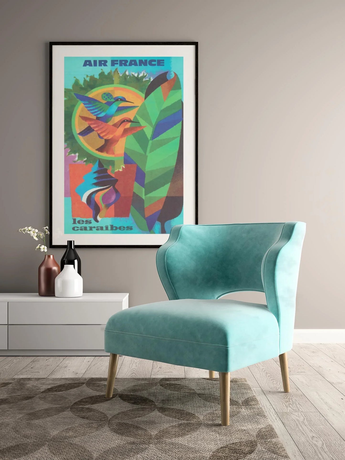 A modern room with a teal velvet chair, a light gray side table holding a white vase with flowers and two bottles, and a colorful Mambí Air France “Les Caraïbes” travel poster on the taupe wall.
