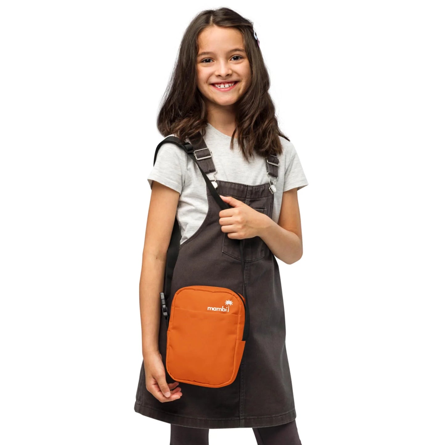 A smiling young girl with long brown hair wears a grey t-shirt, a dark grey overall dress, and an orange Mambí crossbody bag. She stands against a white background.