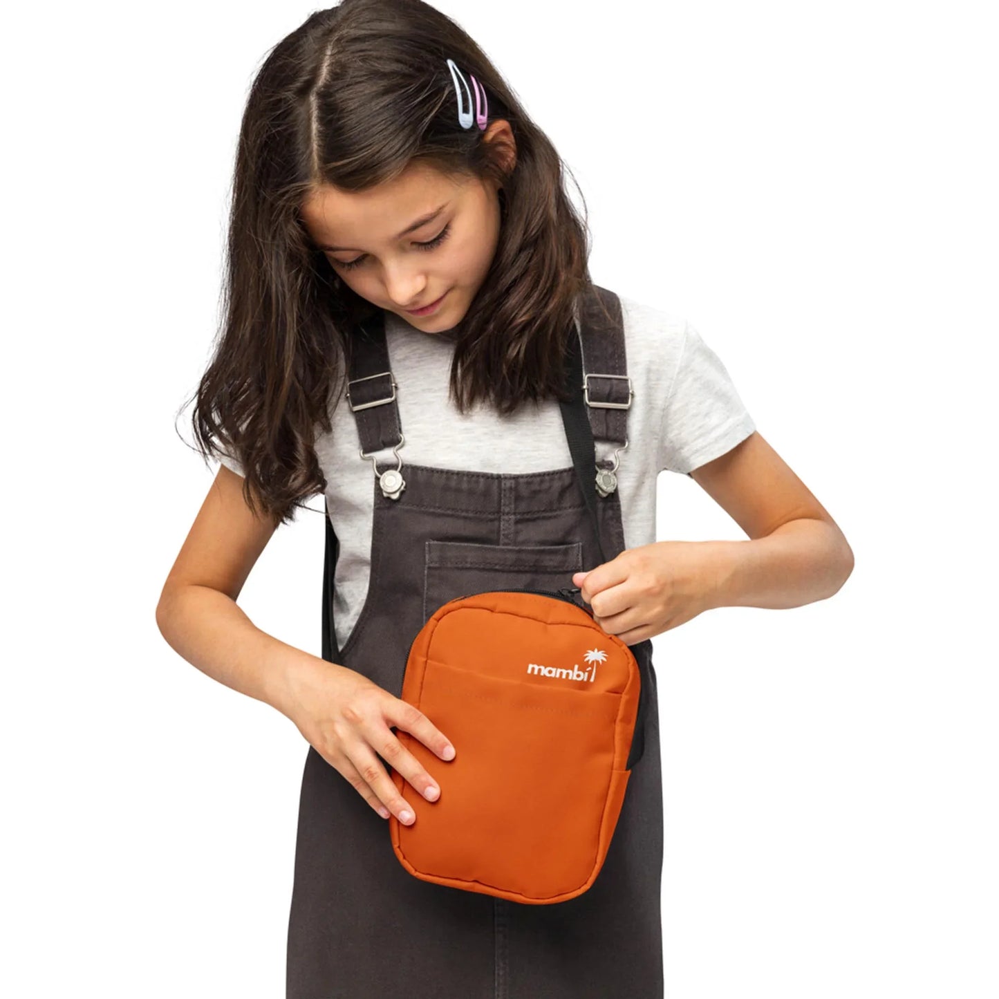 A young girl with dark hair in clips, wearing a grey shirt and dark overalls, looks down while holding and unzipping an orange Mambí lunch bag.