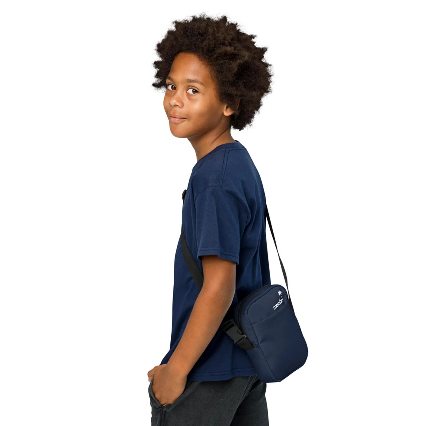 A young person with curly hair, wearing a navy blue Mambí t-shirt and dark pants, stands sideways and smiles slightly while carrying a small navy blue shoulder bag.