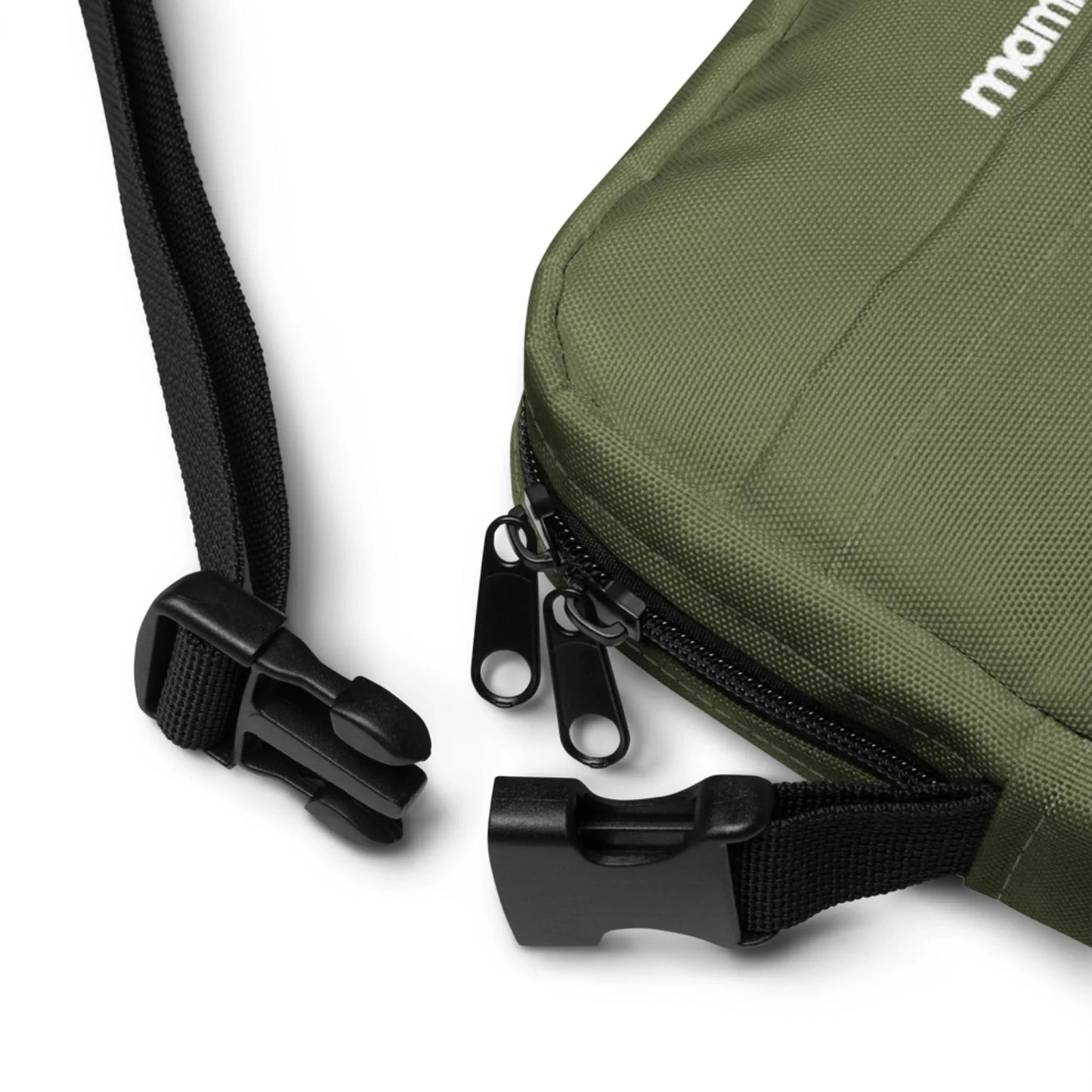 Close-up of a green Mambí fabric bag with black zippers and a black adjustable strap, featuring a plastic buckle clasp. The strap and buckle are unfastened, lying on a white surface.