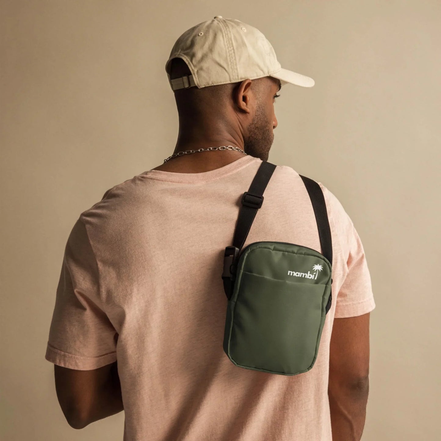 A person wearing a light pink t-shirt and beige cap is seen from behind, carrying a green Mambí crossbody bag against a neutral background.