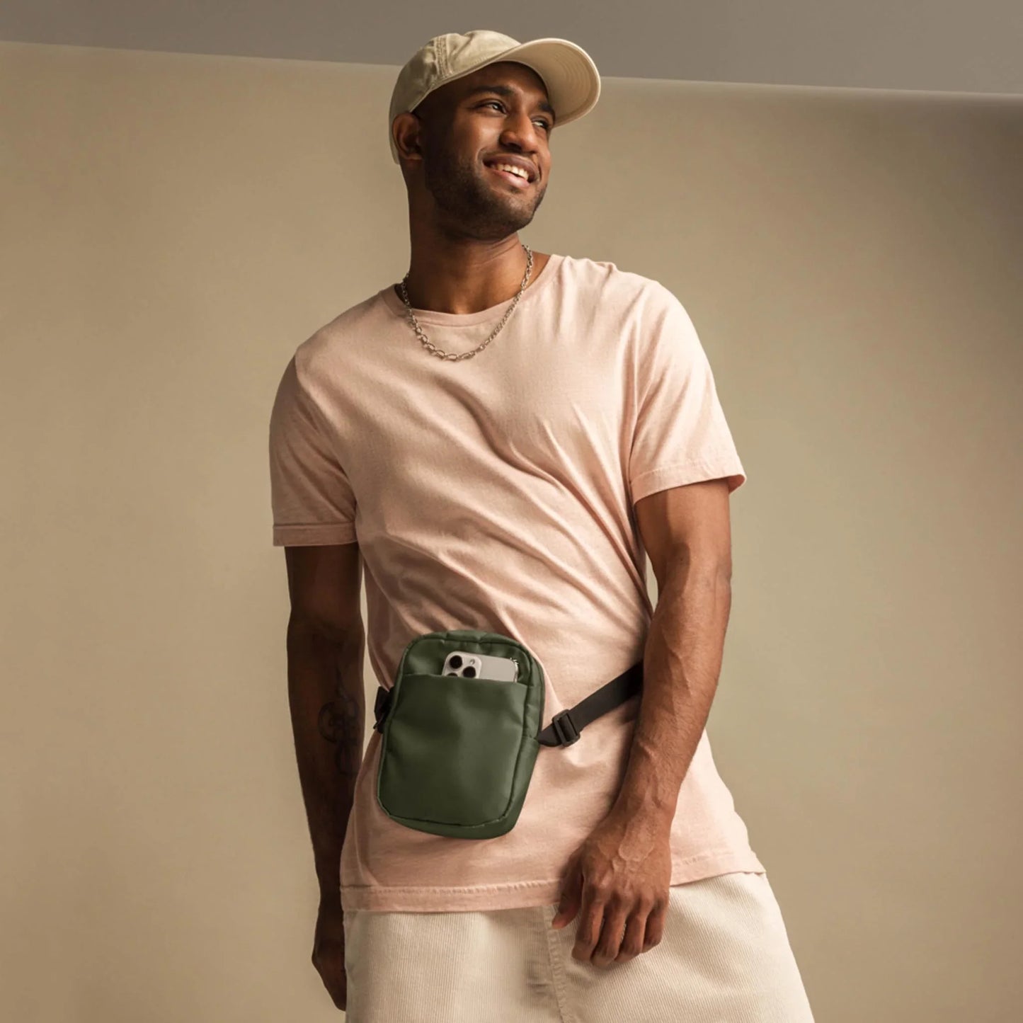 A smiling man in a light pink t-shirt, white shorts, and a beige cap stands against a neutral background. He carries a green Mambí crossbody bag with a phone visible in its front pocket.