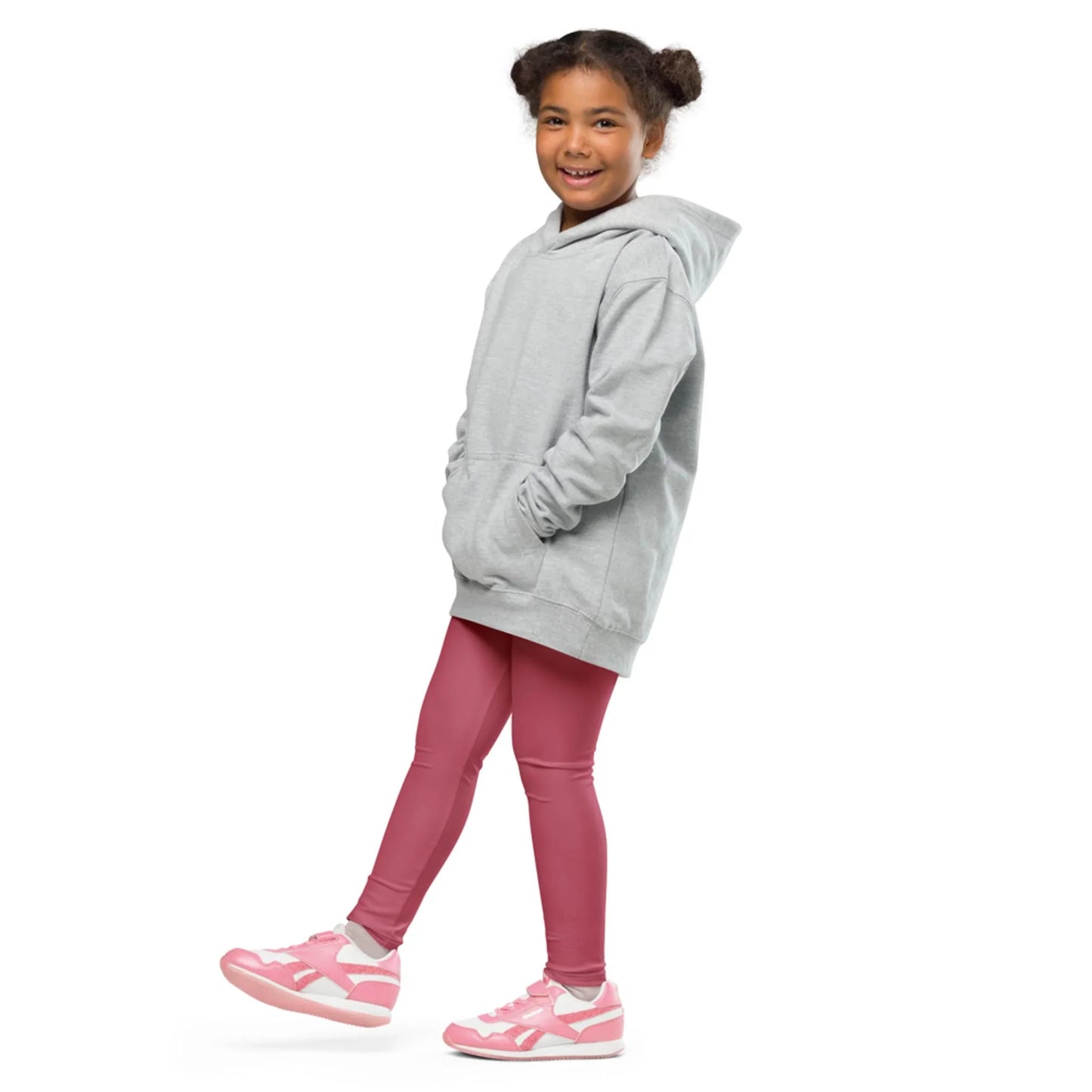 A smiling young girl with curly hair in pigtails wears a gray Mambí hoodie, pink leggings, and pink sneakers. She stands with one foot forward against a white background.