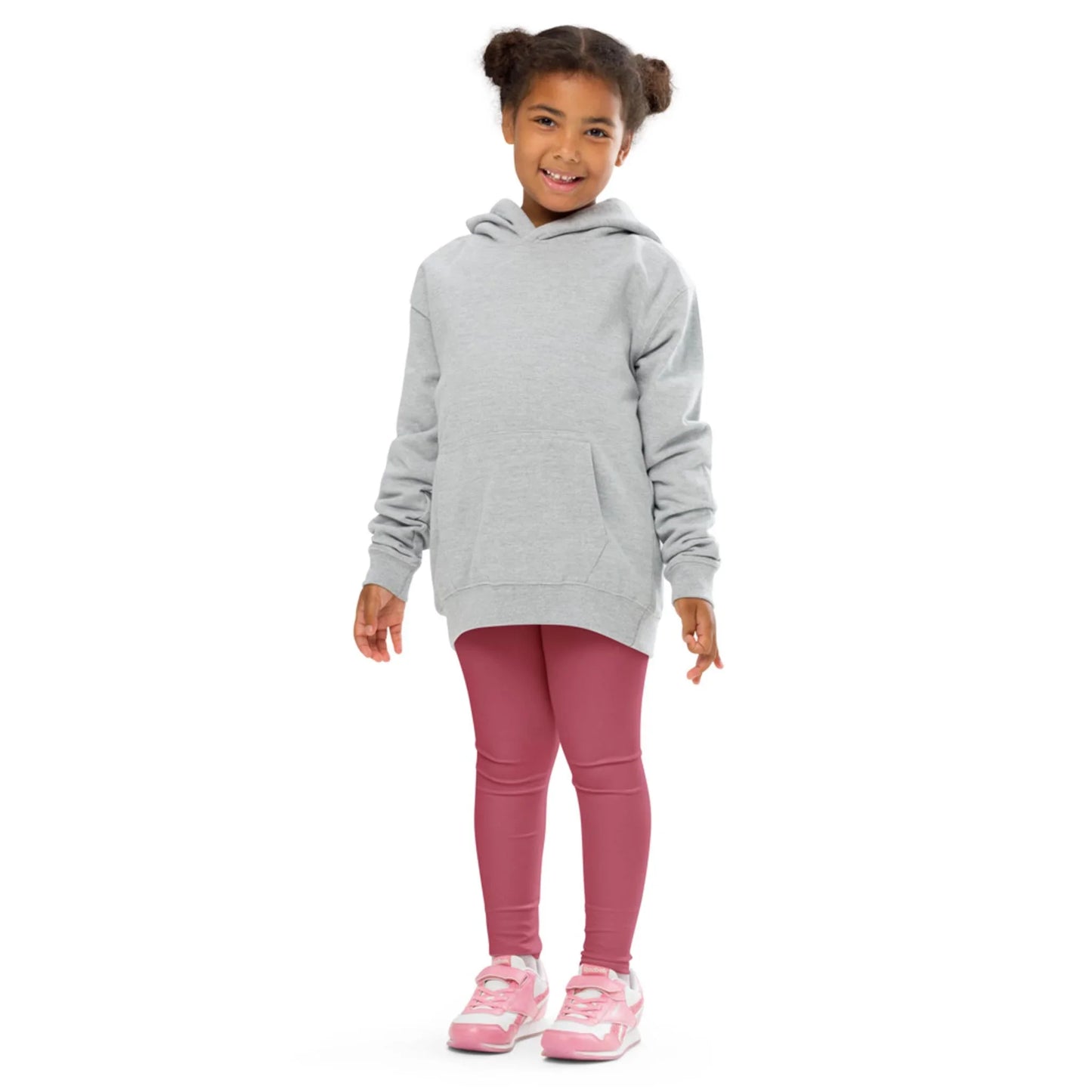 A young girl stands smiling in a light gray Mambí hoodie, pink leggings, and pink sneakers against a plain white background.