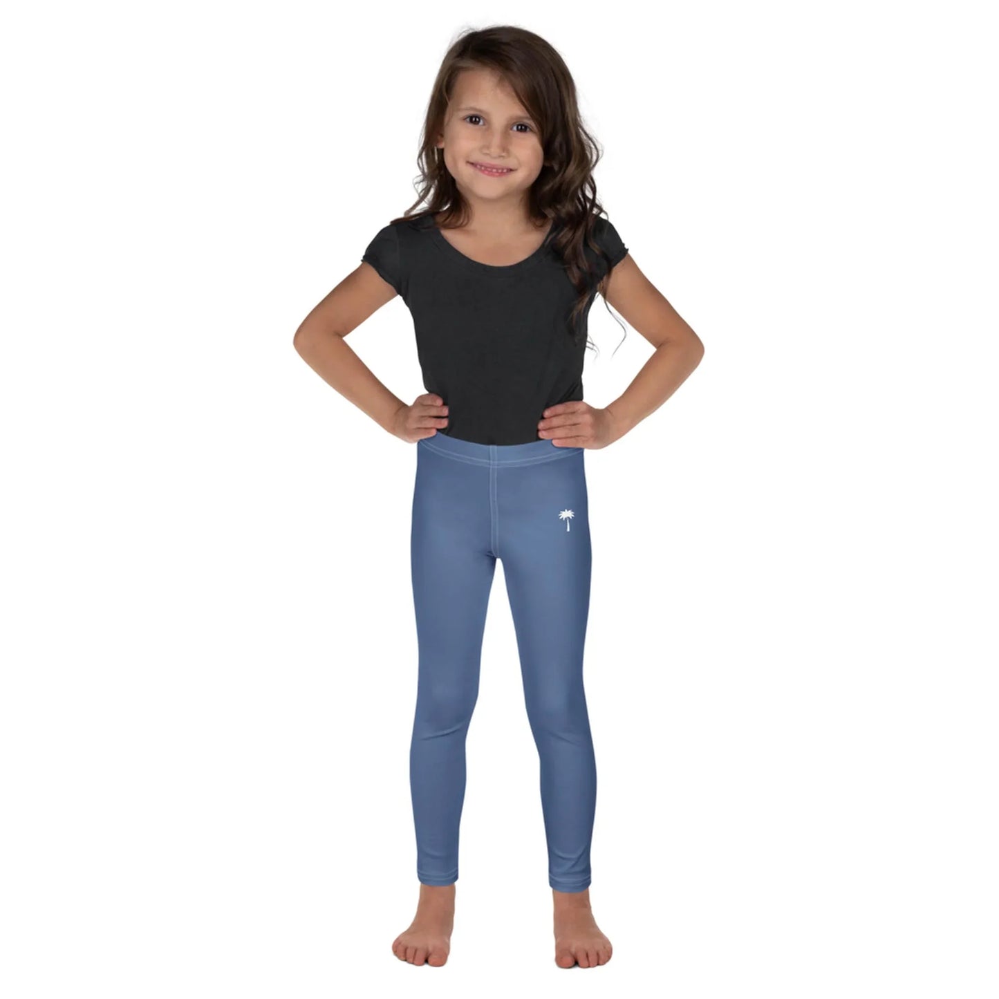 Young girl with long brown hair stands barefoot, smiling with hands on hips. She wears a Mambí black short-sleeve shirt and light blue leggings, standing against a plain white background.